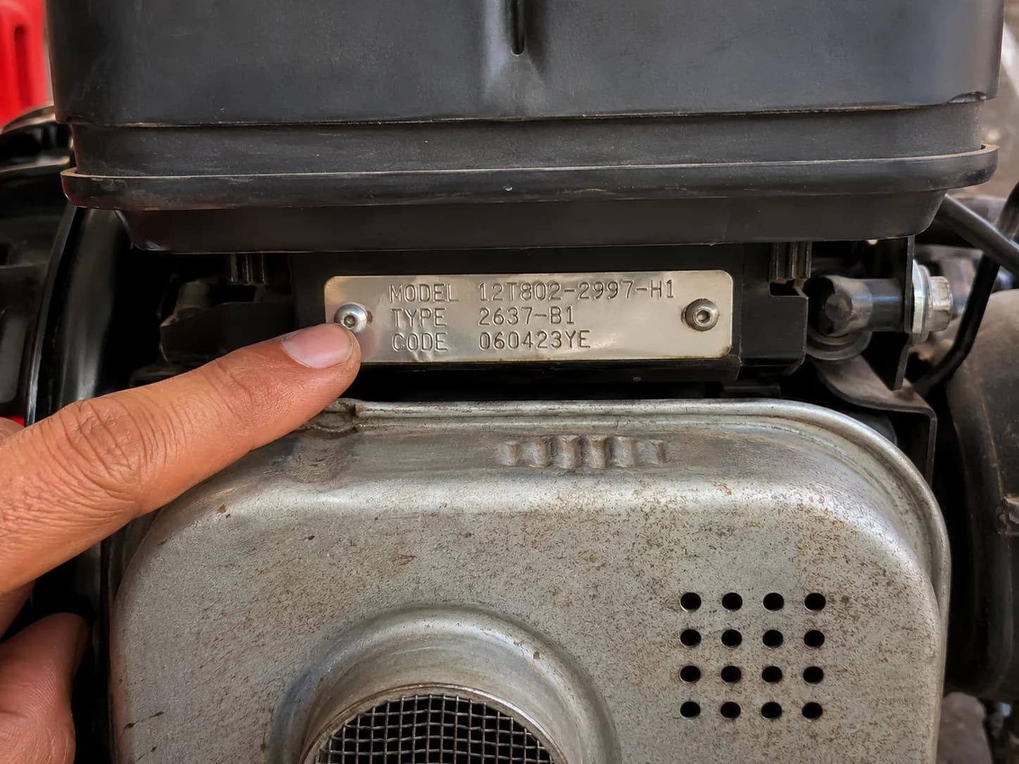 Finger pointing to the Briggs & Stratton engine ID plate above the muffler showing the model, type, and code numbers.