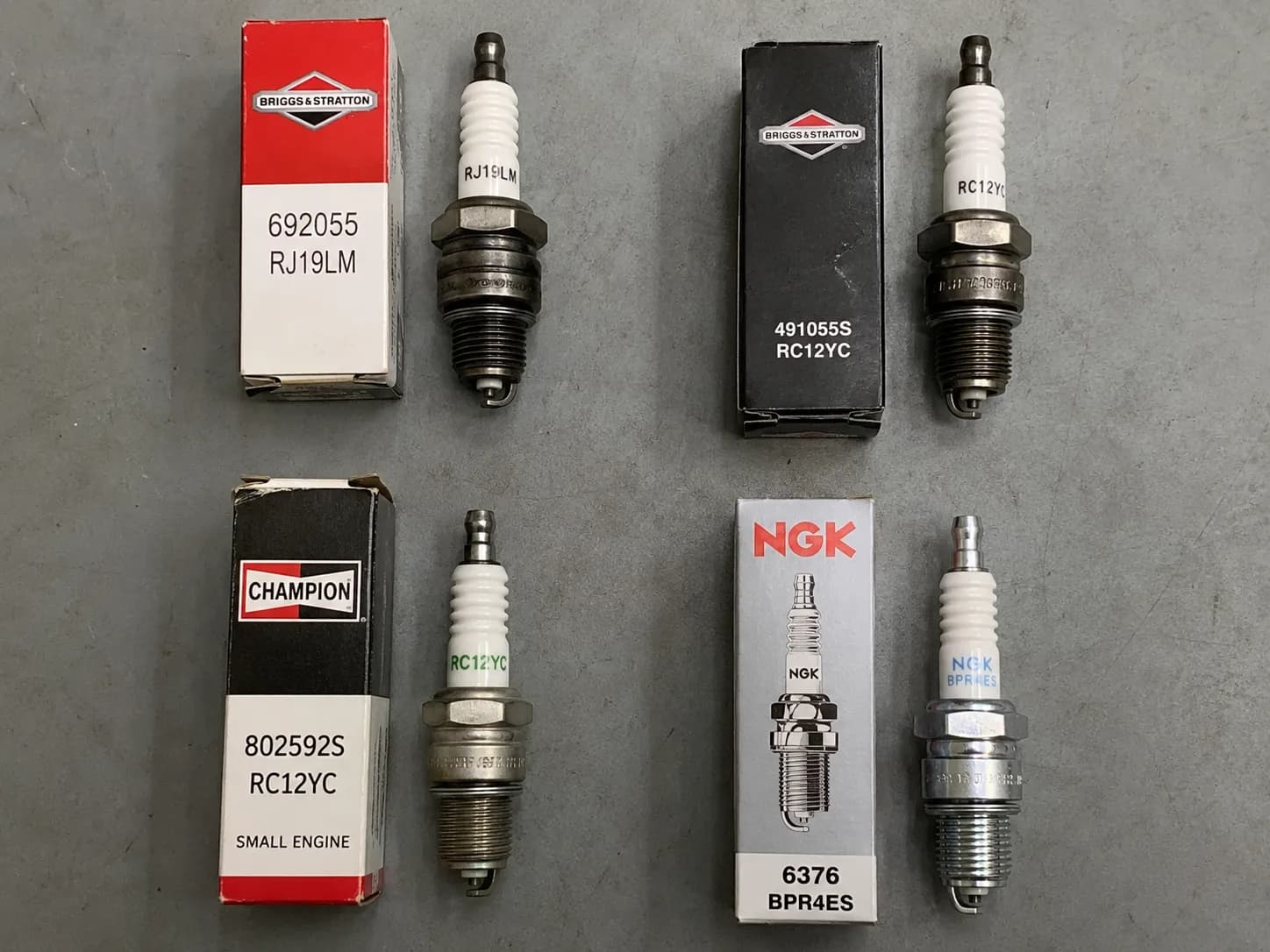Four Briggs & Stratton spark plugs lined up beside their OEM boxes with part numbers visible on a grey workbench.