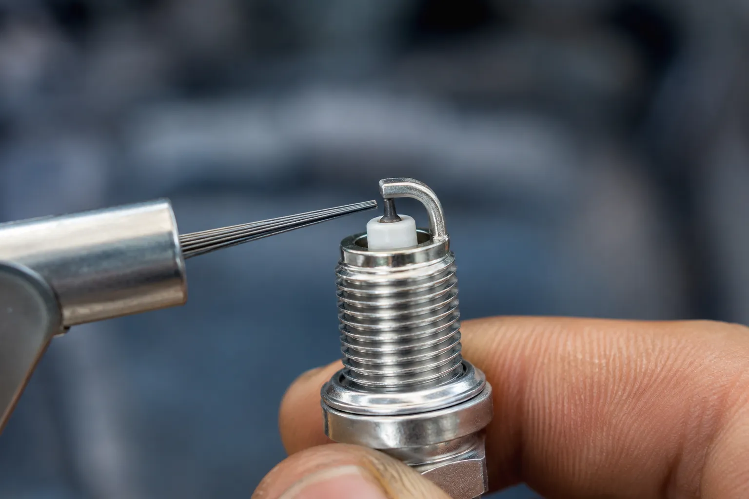 Hand holding a new spark plug while a wire feeler gauge is inserted between the electrodes to check the spark plug gap, with the electrode area in sharp focus.
