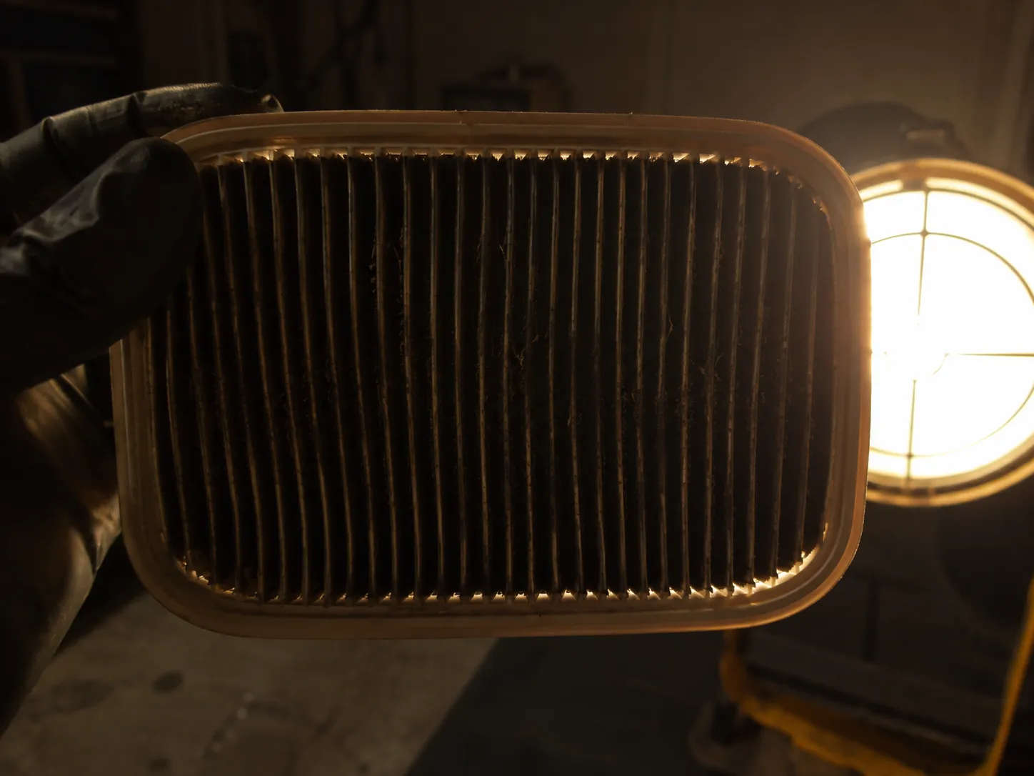 Close-up of a clogged white pleated paper lawn mower air filter held against a bright work light, with dark, dust-filled pleats blocking nearly all light, showing it needs replacement.