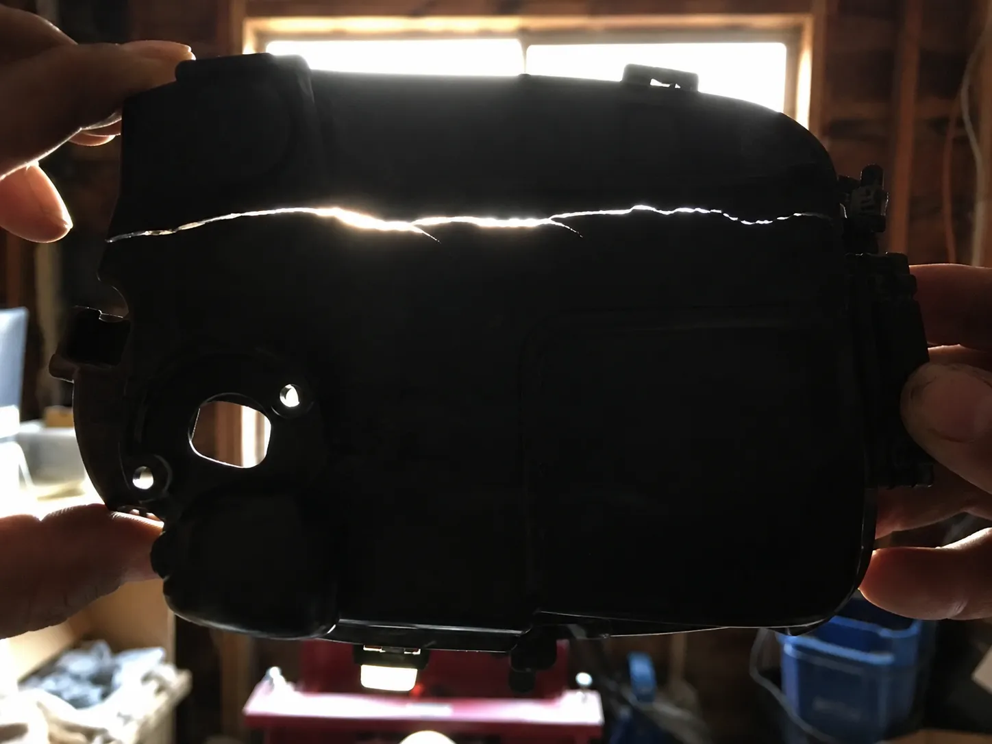 Black plastic lawn mower air-cleaner housing held against a bright window, revealing a thin hairline crack glowing along the molded seam during a backlight inspection.