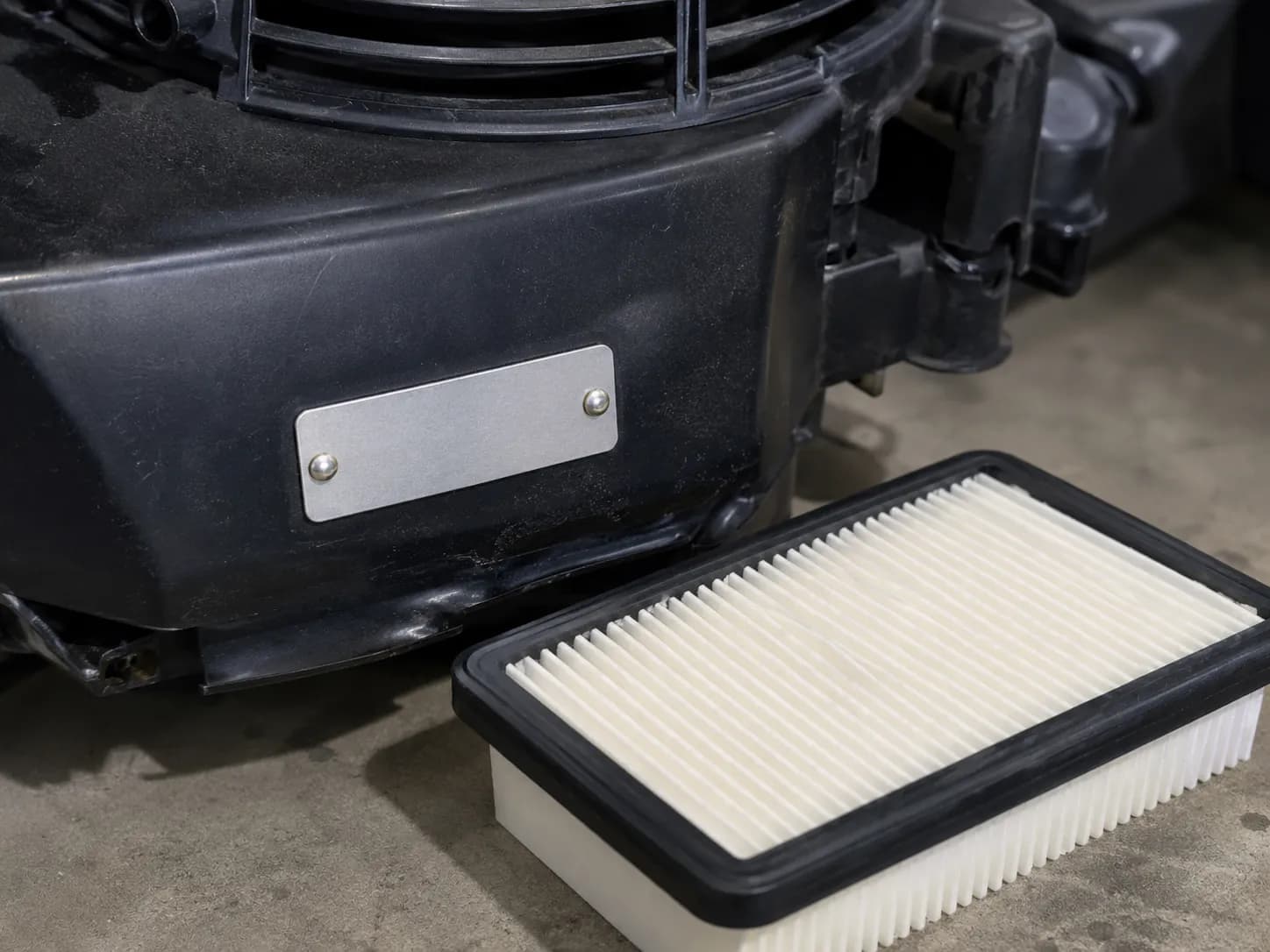Close-up of a small-engine model tag on a black engine shroud beside a white pleated paper air filter on a workbench during routine mower service.