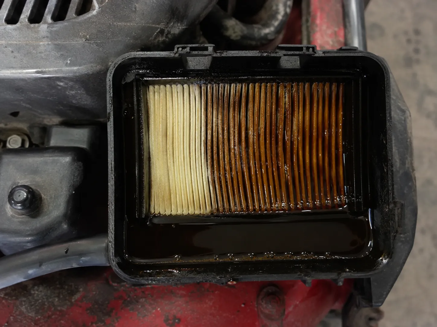 Close-up of engine oil pooled inside a lawn mower air filter housing after the mower was tipped incorrectly, showing a paper air filter soaked with oil along one edge.