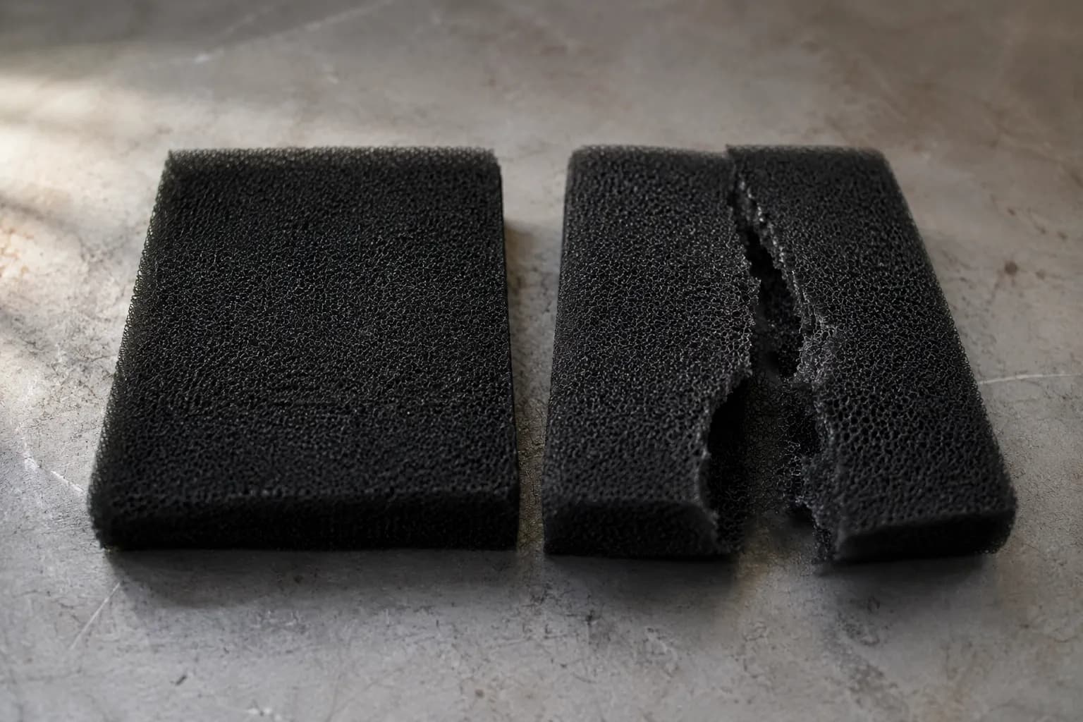 Two black foam lawn mower air filters on a gray workbench, showing an intact filter beside a torn filter split along a fold after wringing, highlighting proper squeeze-only cleaning.