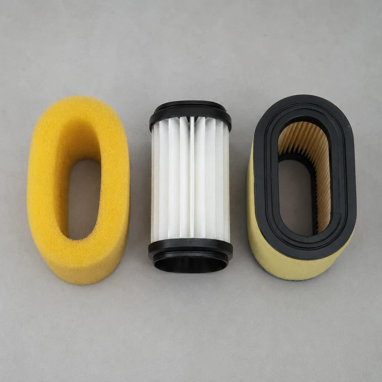 Side-by-side comparison of foam, pleated paper, and dual-element lawn mower air filters on a light grey background, showing the three common air filter types used in small engines.