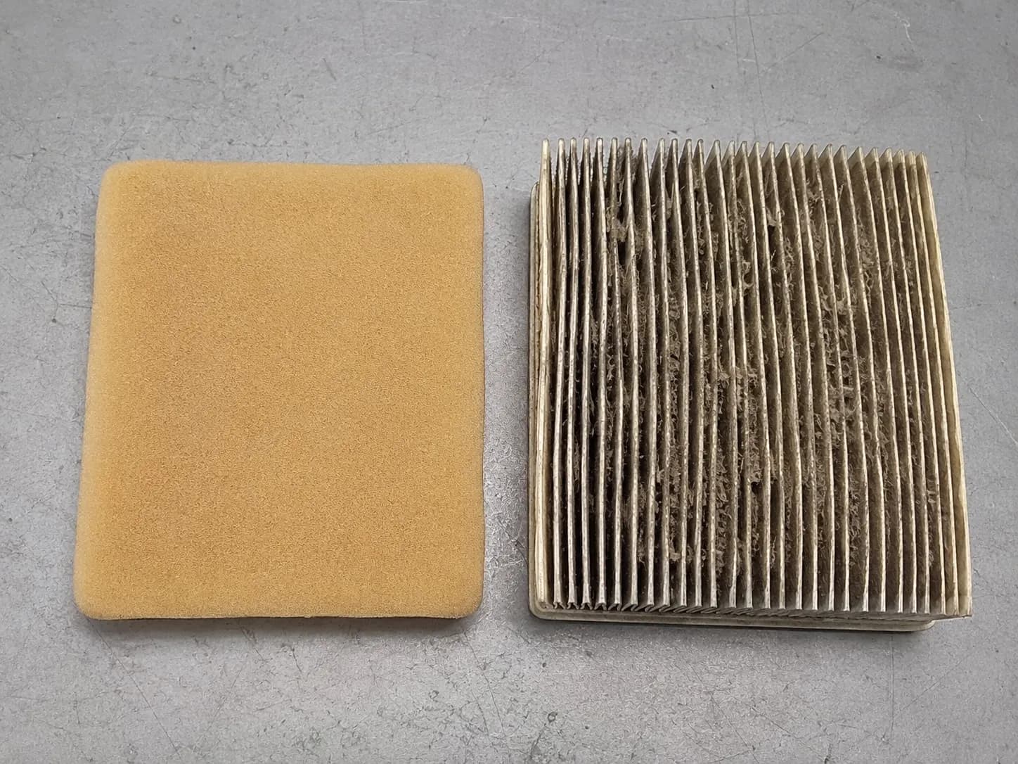 Clean tan foam pre-cleaner beside a dirty grey pleated paper lawn mower air filter with dust-clogged folds, showing why paper elements are replaced instead of washed.