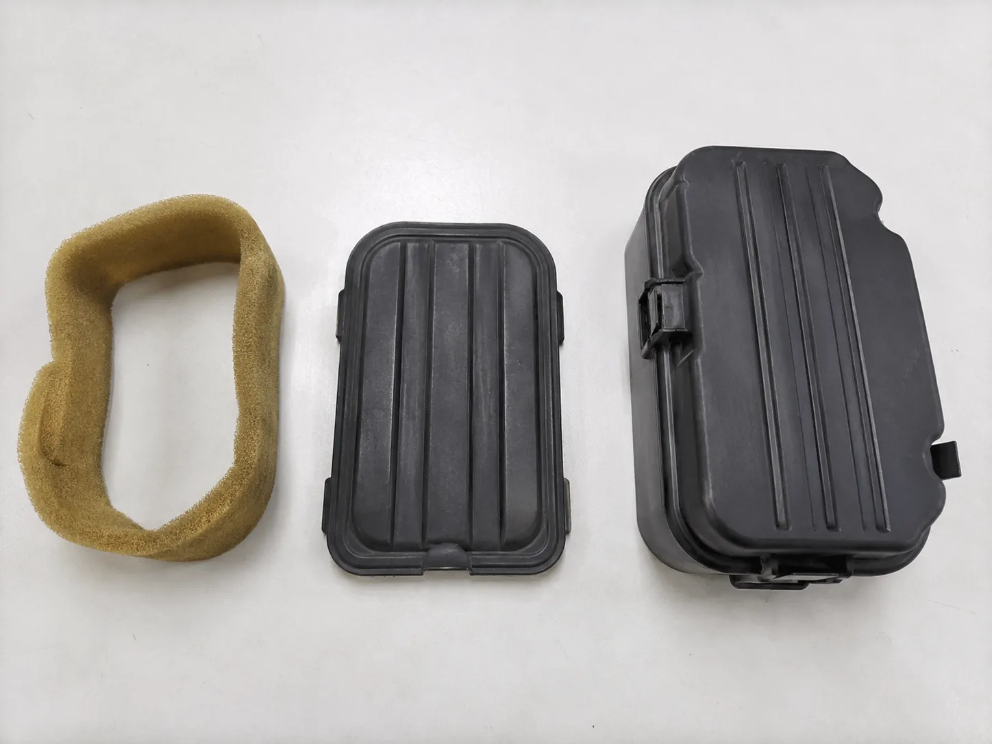 Foam pre-cleaner sleeve beside two Honda-style small engine air filter covers, showing an older flat design and a newer taller housing on a clean light surface.