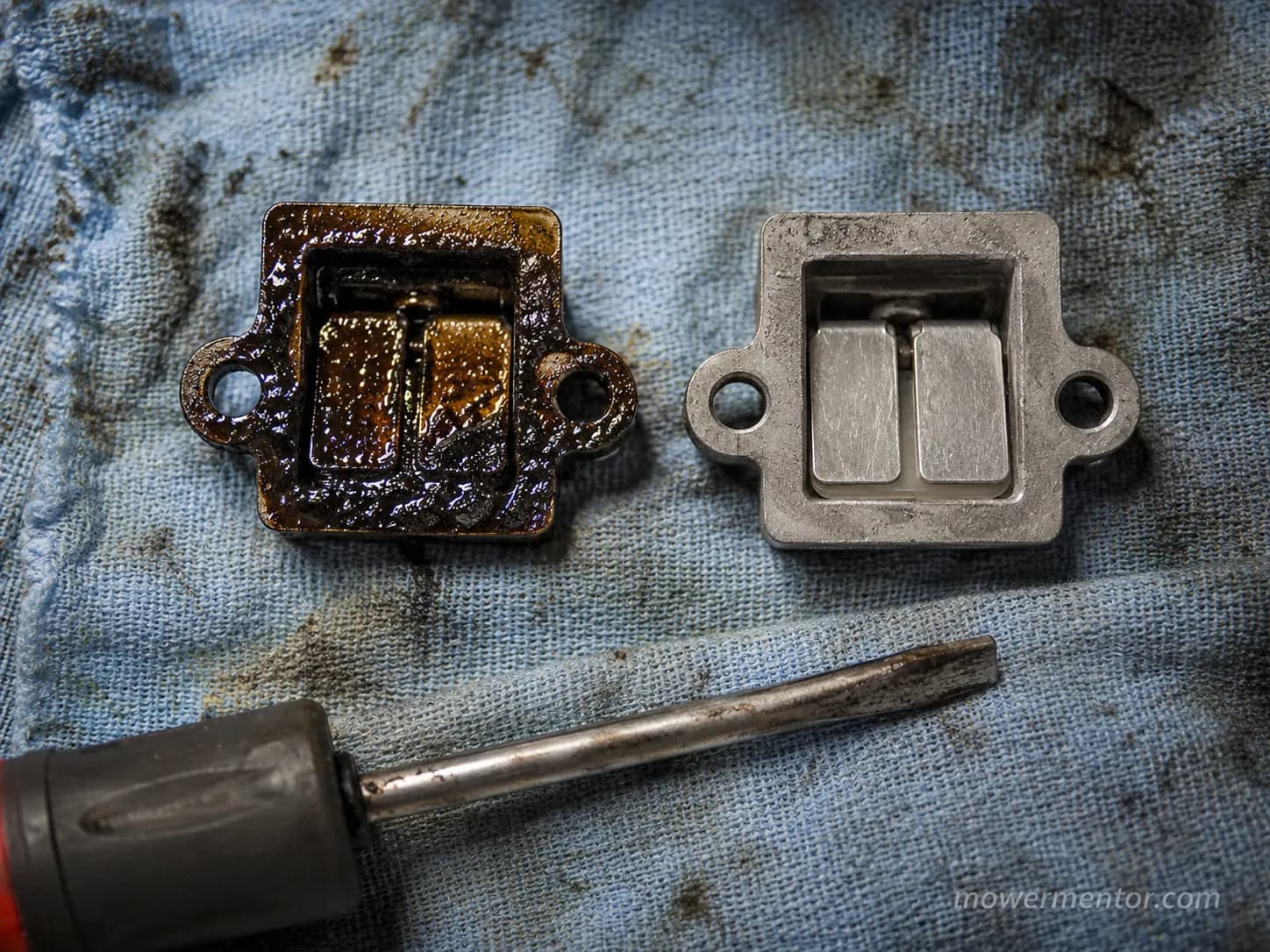 Gummed crankcase breather assembly beside a clean one on a blue shop rag, showing dark oil varnish buildup and clean metal passages during mower engine repair.