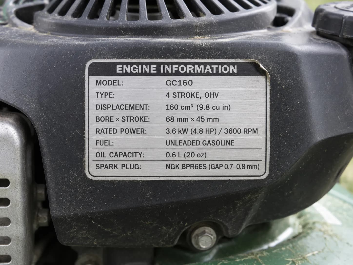 Close-up of a lawn mower engine identification sticker on the blower housing, showing a weathered silver and black decal with model information, light grass dust, and outdoor daylight.