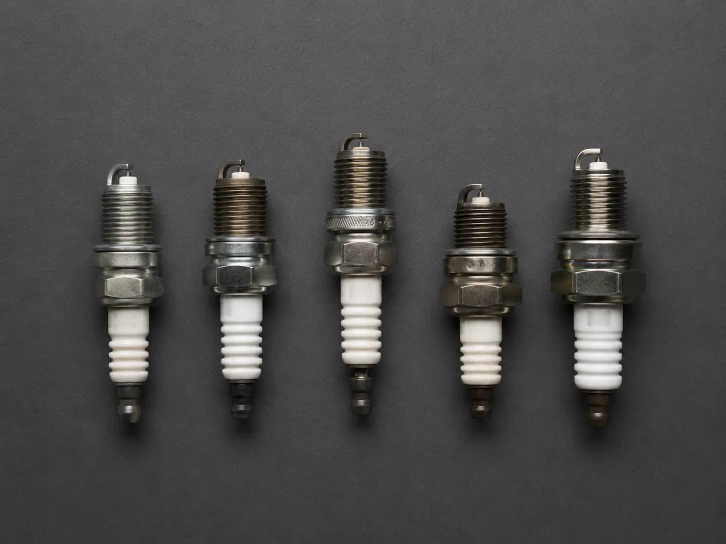 Five lawn mower engine spark plugs arranged in a row, showing differences in thread reach, hex size, and overall length across common small engine spark plug types.