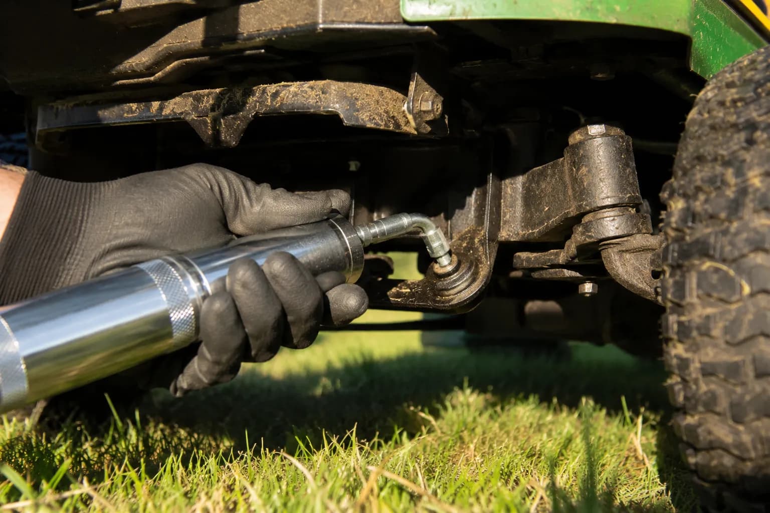 Gloved hand using a grease gun to lubricate a zerk fitting on the front axle pivot of a riding lawn mower, photographed from ground level during routine mower maintenance.