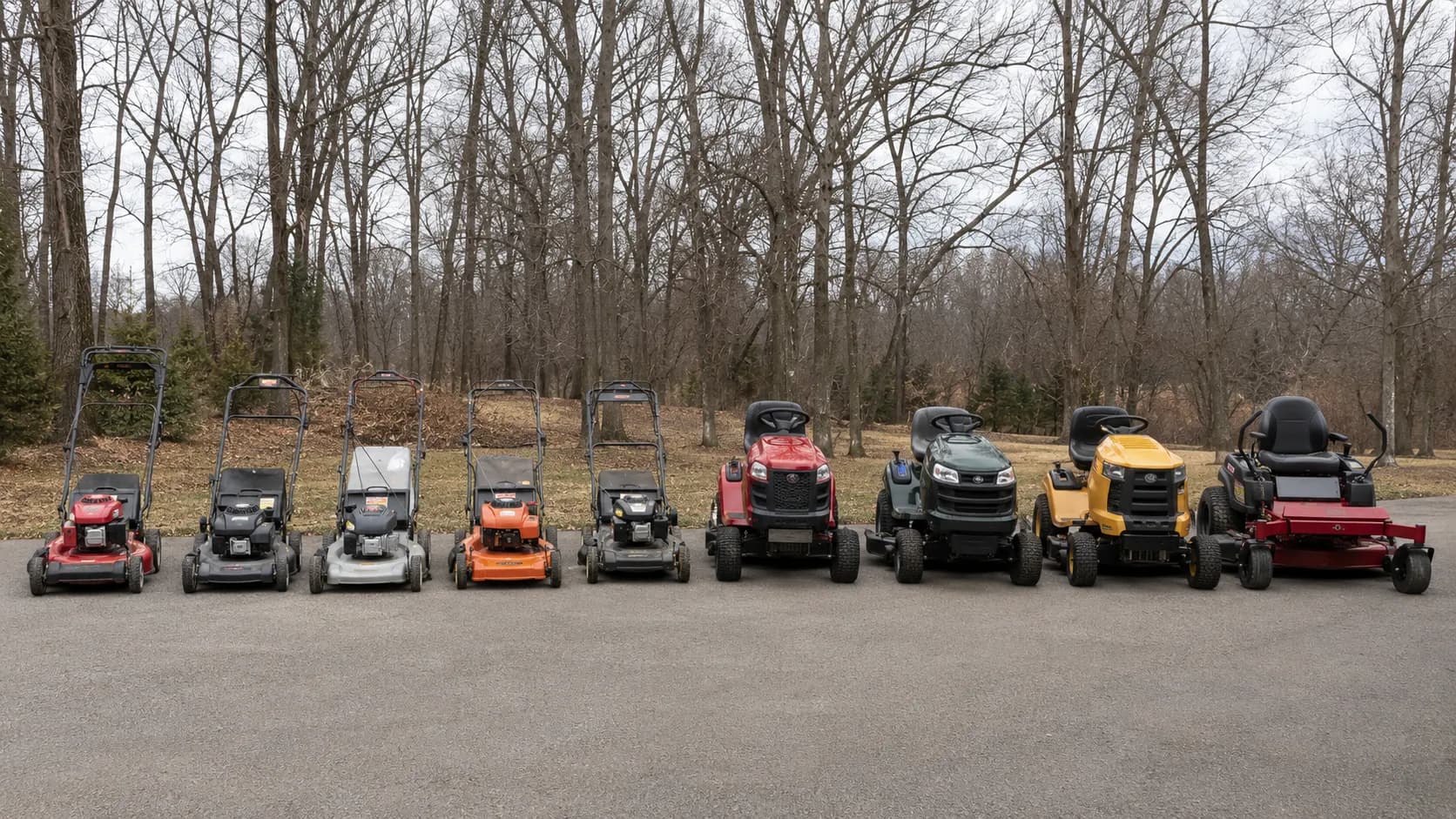 Nine gas lawn mowers lined up on a driveway for testing, with five walk-behind and four riding mowers photographed in early spring under overcast skies on 3 March 2026.