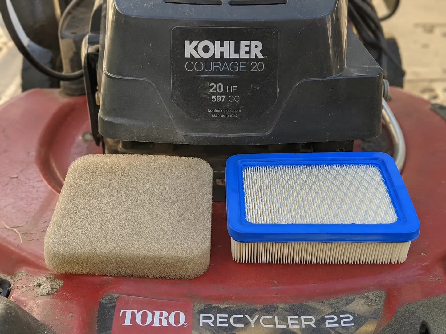 Kohler Courage engine shroud label on a Toro Recycler mower with a tan foam pre-cleaner and a blue-edged pleated paper air filter resting on the mower deck.