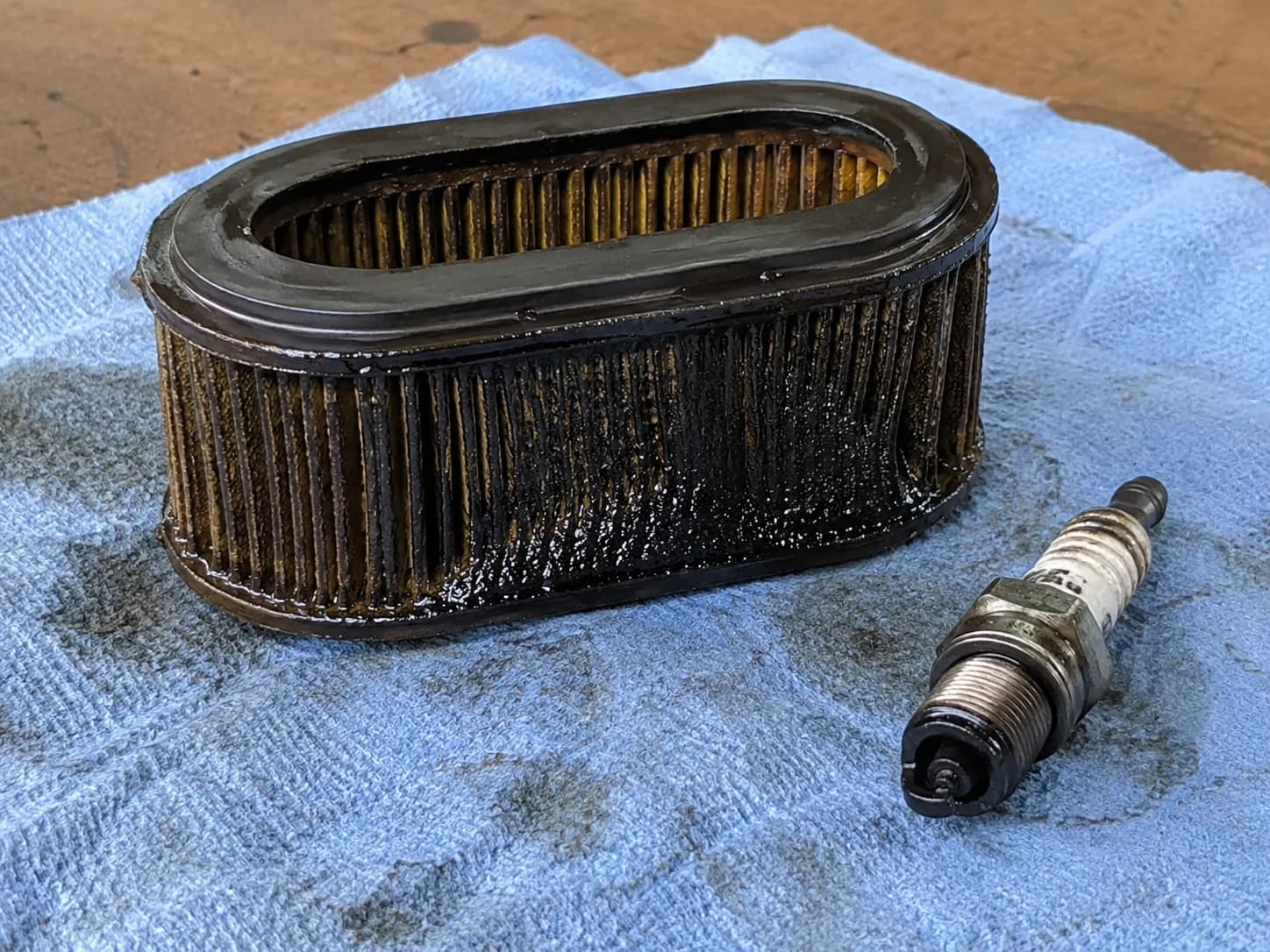 Oil-soaked mower paper air filter beside a fouled spark plug on a blue shop rag after the mower was tipped the wrong way, showing oil contamination and ignition fouling.