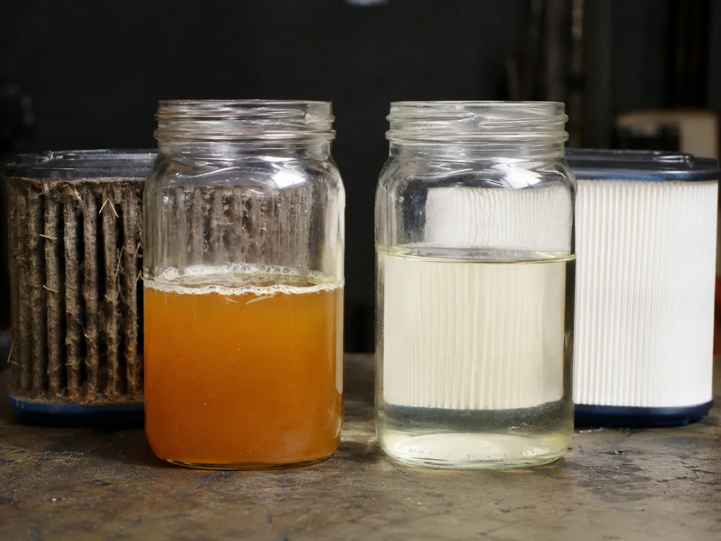 Side-by-side comparison of old varnished mower fuel and fresh gasoline in glass jars, with dirty and clean air filter elements behind, showing how stale fuel affects small engine performance.