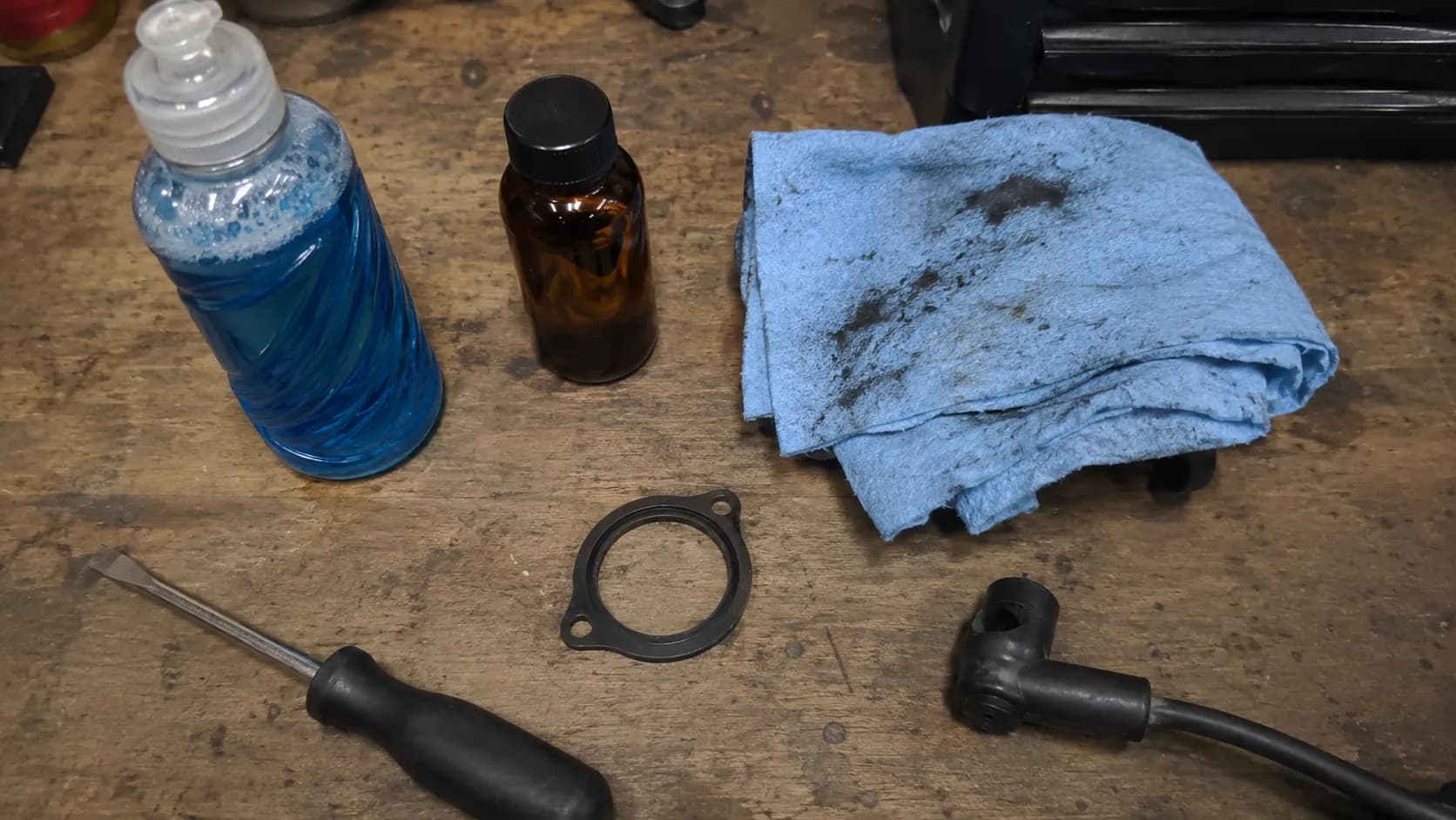 Top-down view of a small-engine service workbench with dish soap, engine oil, a shop rag, spare air-cleaner gasket, flat-head screwdriver, and a disconnected spark-plug boot.