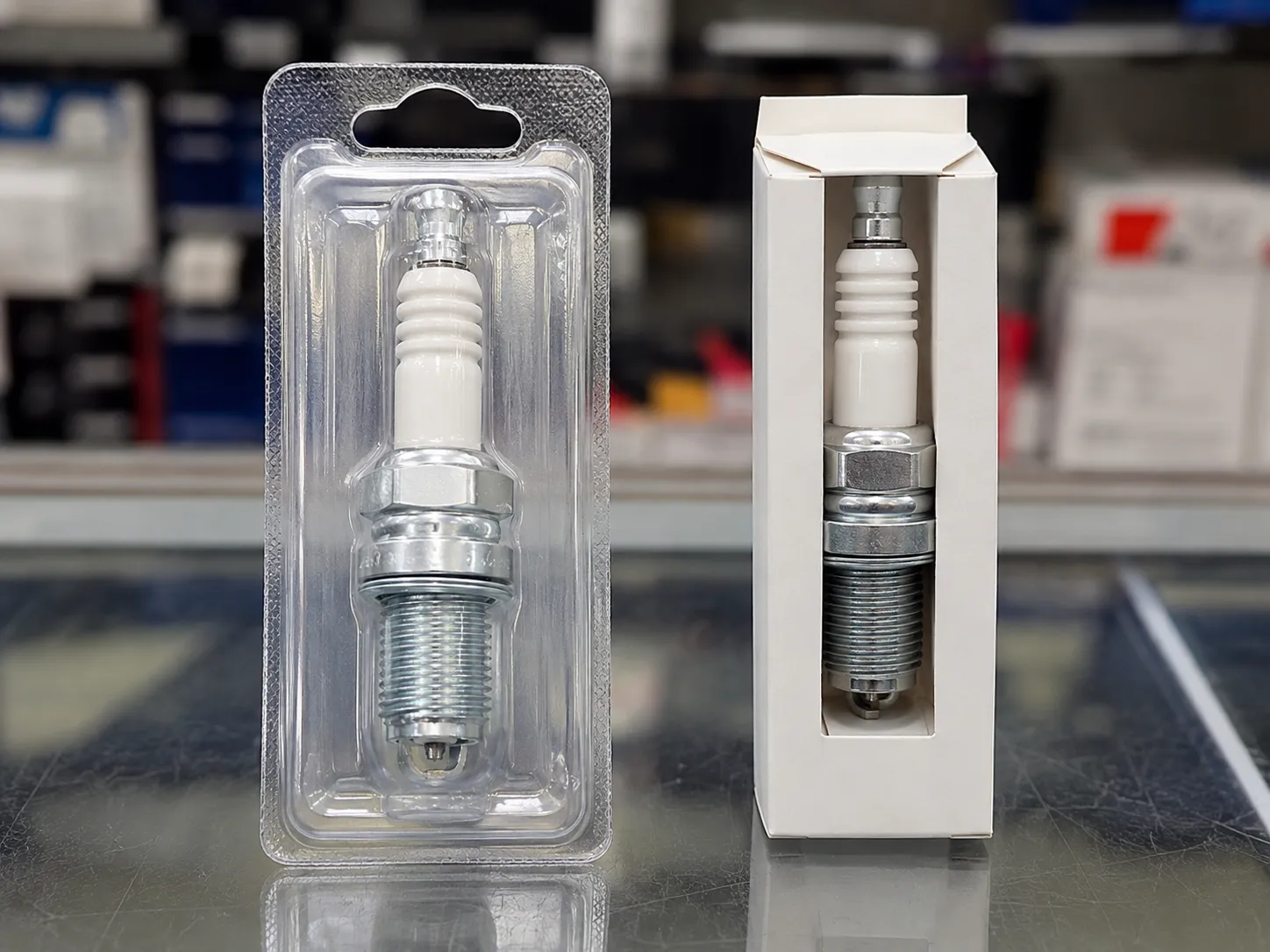 A spark plug displayed in plain aftermarket blister packaging beside the same style of spark plug in a plain OEM-style box on a retail counter for packaging comparison.