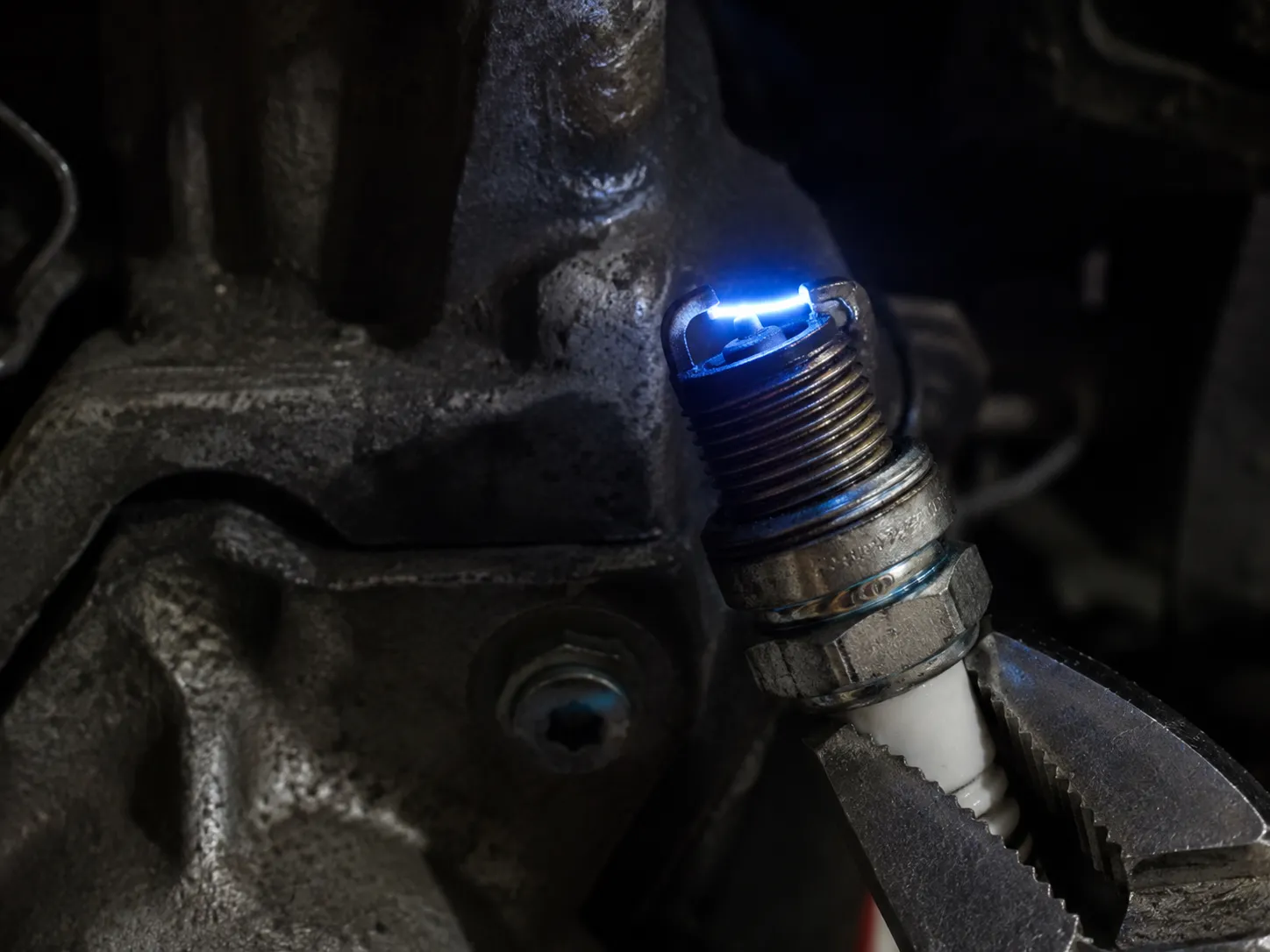 Close-up of a small engine spark plug held with insulated pliers as a bright blue spark jumps across the electrode gap against the engine block during an ignition spark test.