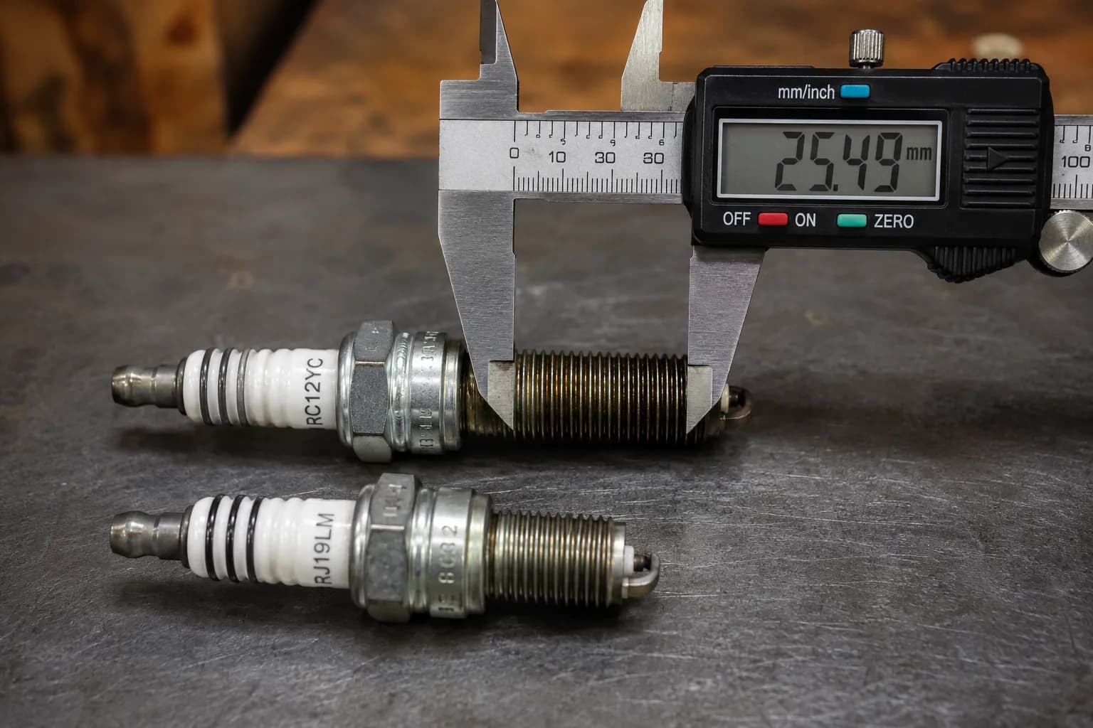 Two small-engine spark plugs side by side with digital calipers measuring the longer plug's threaded reach to compare thread length differences.