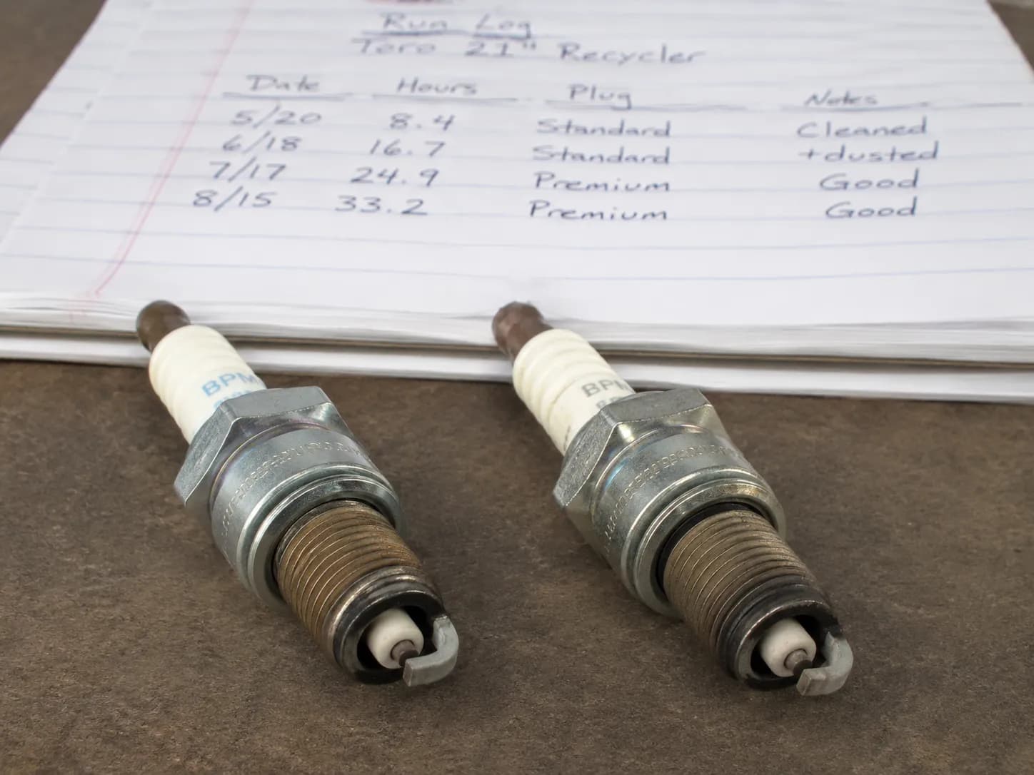 Standard and premium small-engine spark plugs after one season of use beside a handwritten maintenance log, showing light electrode wear for comparison.