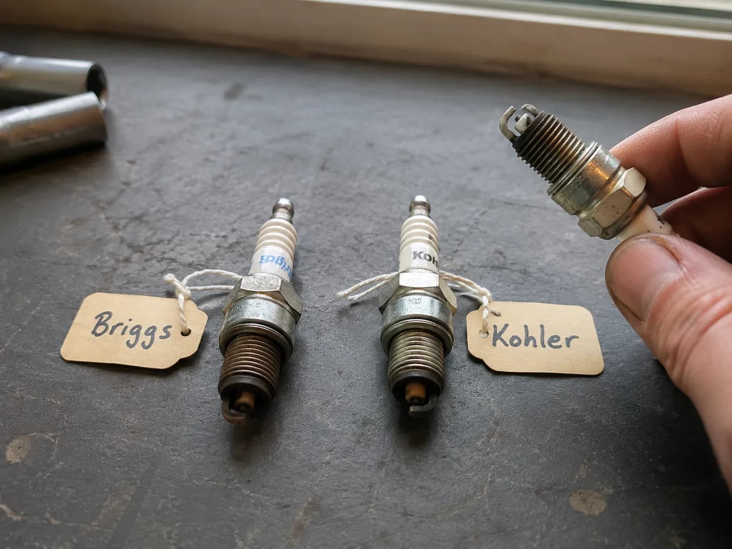Two used Toro lawn mower spark plugs labeled Briggs and Kohler lie side by side on a workbench while a hand holds a third spark plug for comparison.