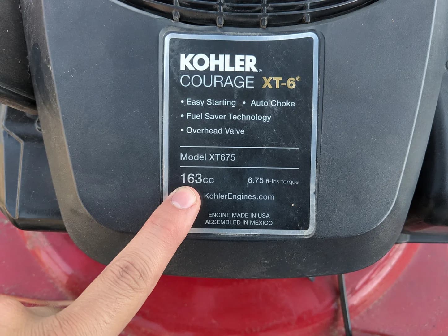 Finger pointing to the engine shroud sticker on a Toro lawn mower, highlighting the manufacturer and engine displacement information for model identification.