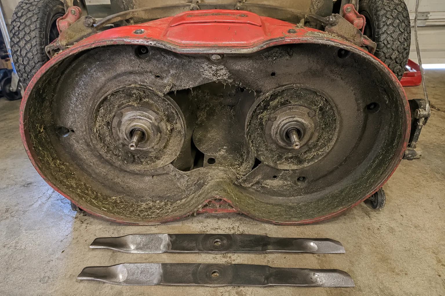 Underside of a Toro TimeMaster mower deck during maintenance, showing the twin-blade spindle assembly with both blades removed and placed on the garage floor for sharpening.