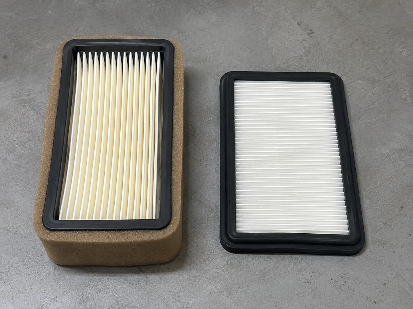 Side-by-side comparison of a deep pleated Toro air filter with tan foam wrap and a thinner Briggs flat panel filter on a gray workbench, highlighting their different designs.