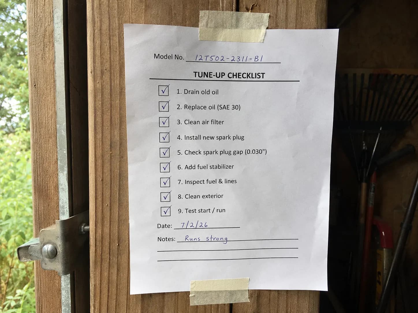 Printed lawn mower tune-up checklist taped inside a wooden shed door with completed checkboxes and a handwritten engine model number at the top.