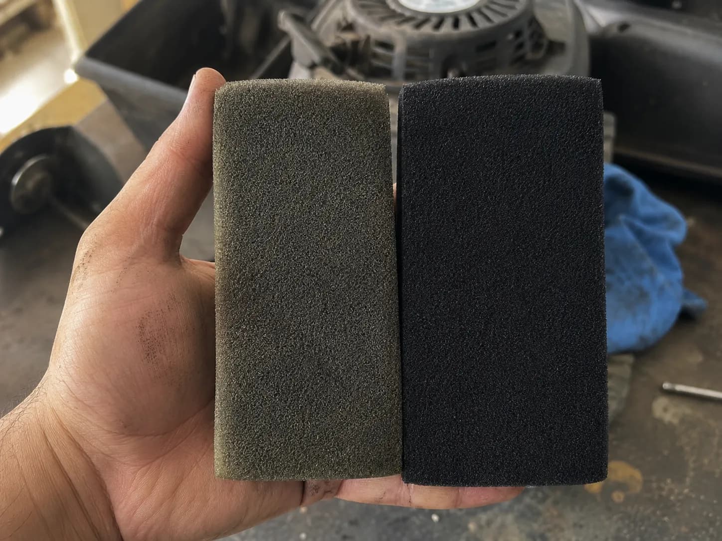 Hand holding a used grey-green foam mower air filter beside a new black foam filter to show that color alone does not indicate whether a foam air filter needs replacement.