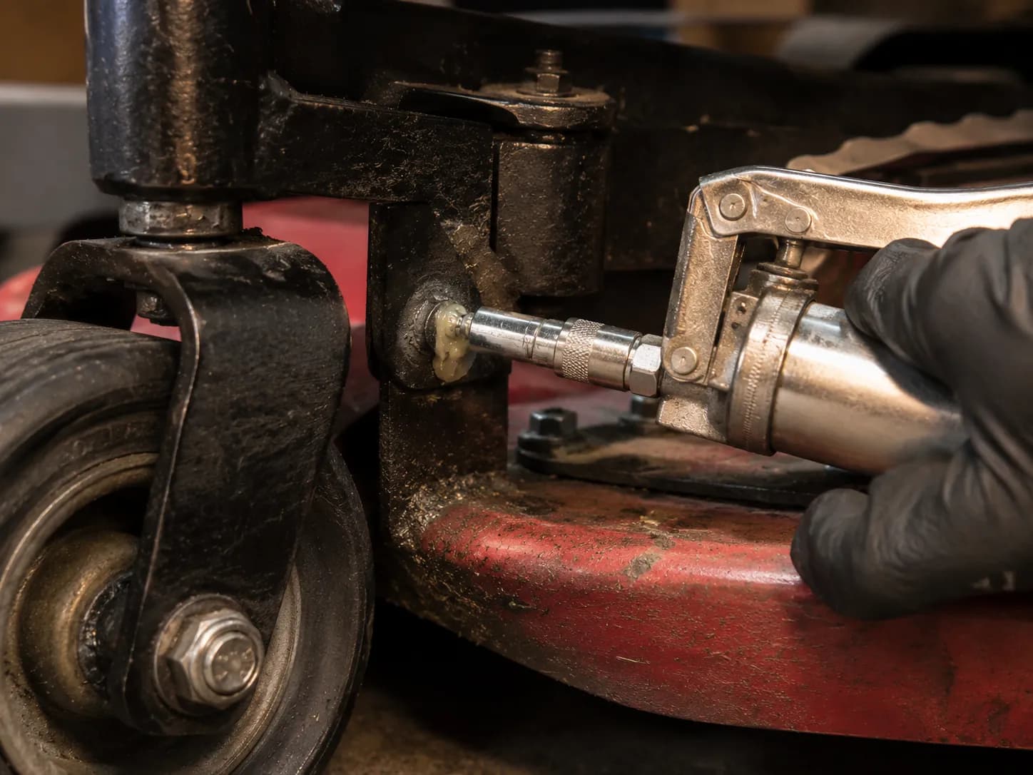 Close-up of a grease gun lubricating a zero-turn mower deck grease fitting, with fresh grease appearing at the zerk fitting while a gloved hand services the mower suspension pivot.