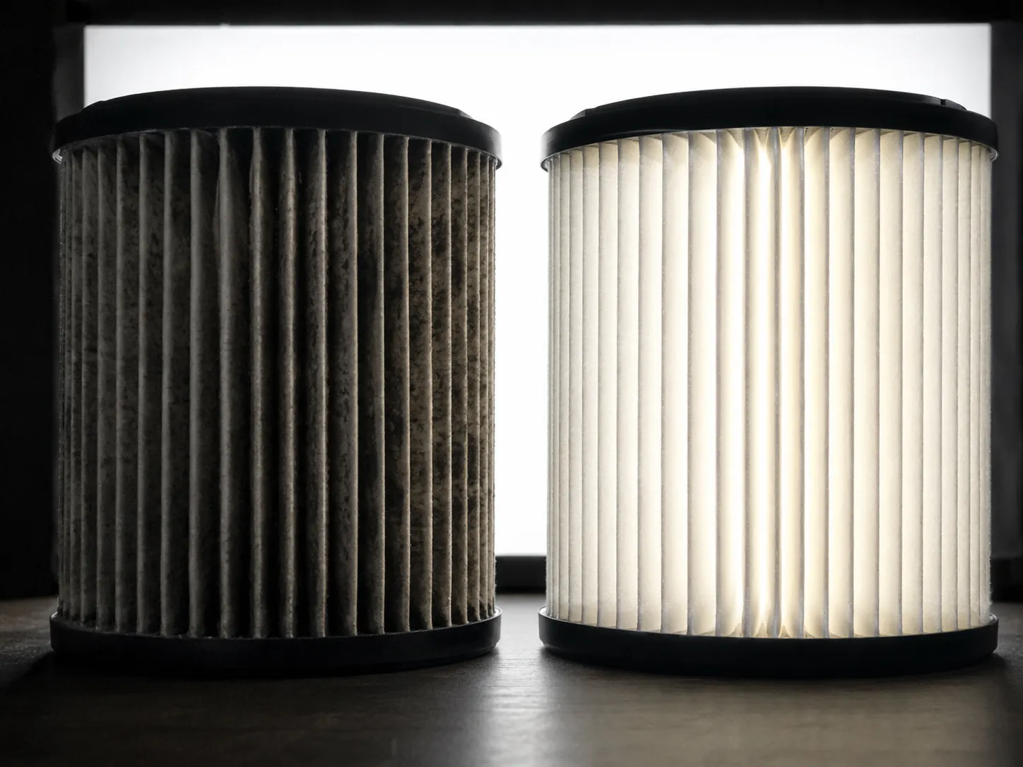 Backlit comparison of a dirty and clean cylindrical paper air filter. The clogged gray filter blocks most light, while the clean white filter glows evenly through its pleated media.