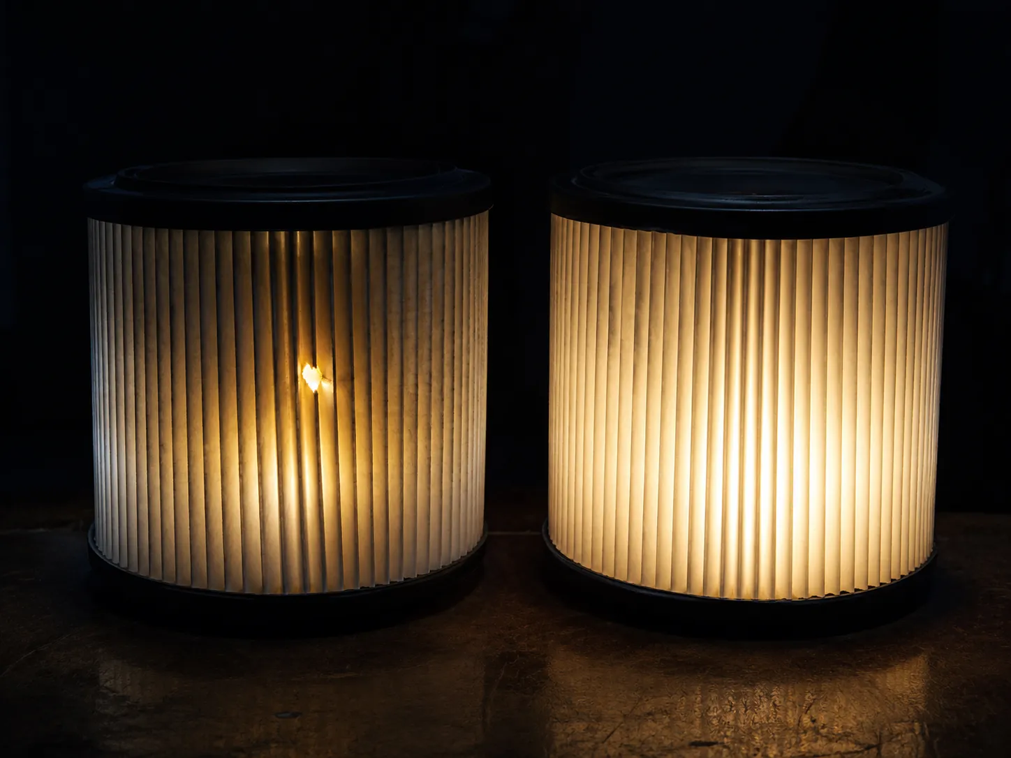 Two pleated paper air filters illuminated from inside in a dark room, with one filter showing a bright pinhole of light indicating damage while the other glows evenly without any leaks.