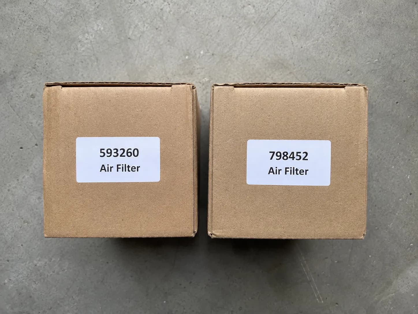Two boxed small engine air filter reference parts placed side by side on a concrete surface with part number labels facing the camera for easy comparison during replacement.