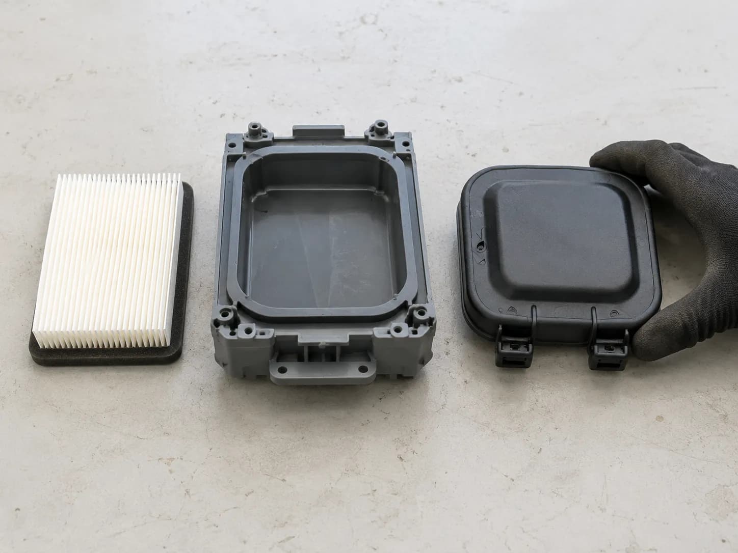 Overhead view of Briggs 550EX lawn mower air cleaner parts showing the black air filter cover, grey air cleaner base, and white pleated filter element arranged side by side.