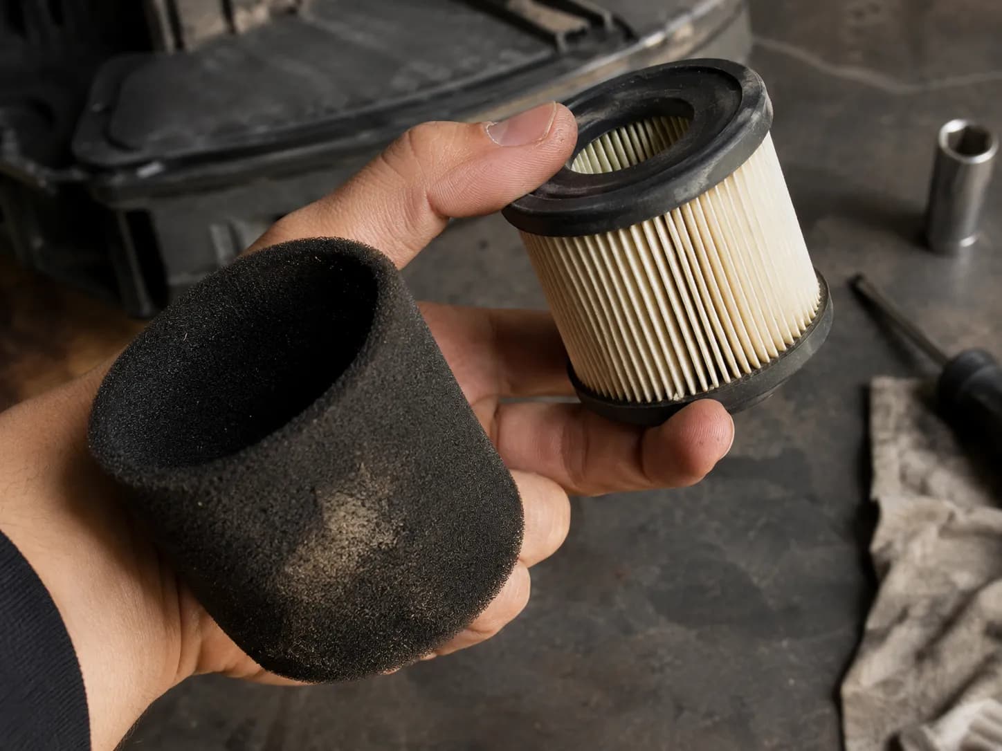 Hand holding a Briggs-style dual-element air filter separated into a black foam pre-cleaner sleeve and a pleated paper cartridge over a workshop bench.