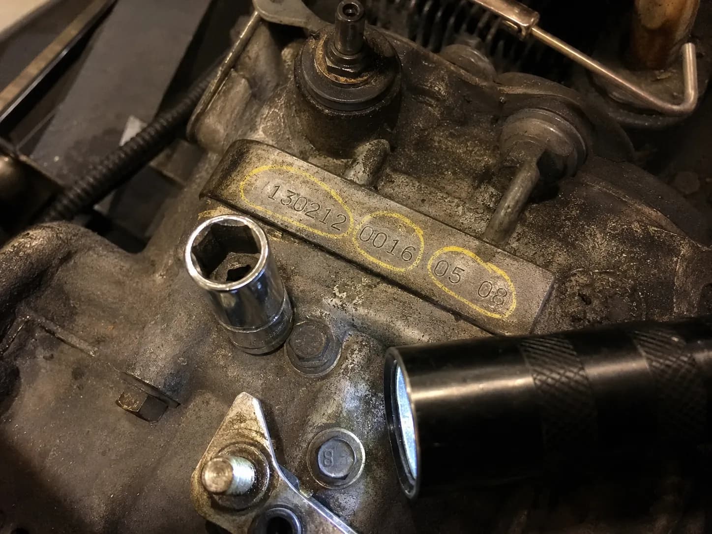 Close-up of a Briggs engine identification stamp with the model, type, and code numbers circled in grease pencil after lifting the engine shroud, with a socket and flashlight nearby.