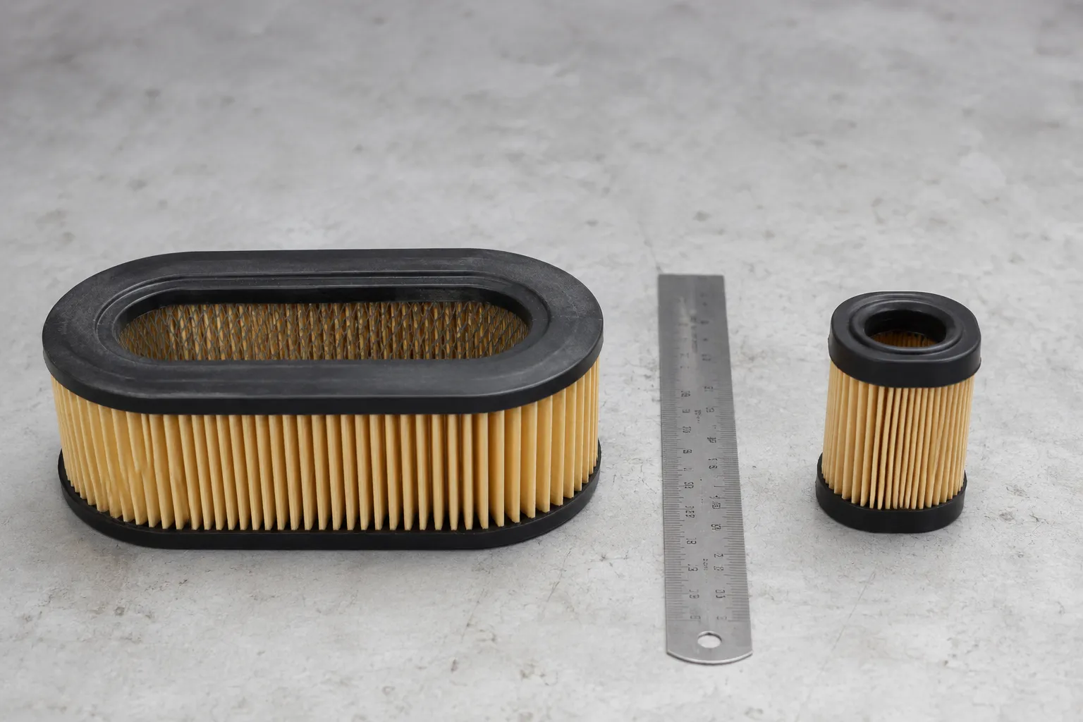 Briggs M110 flat oval paper air filter beside a smaller Tecumseh pleated air filter with a ruler showing the clear difference in size and shape.