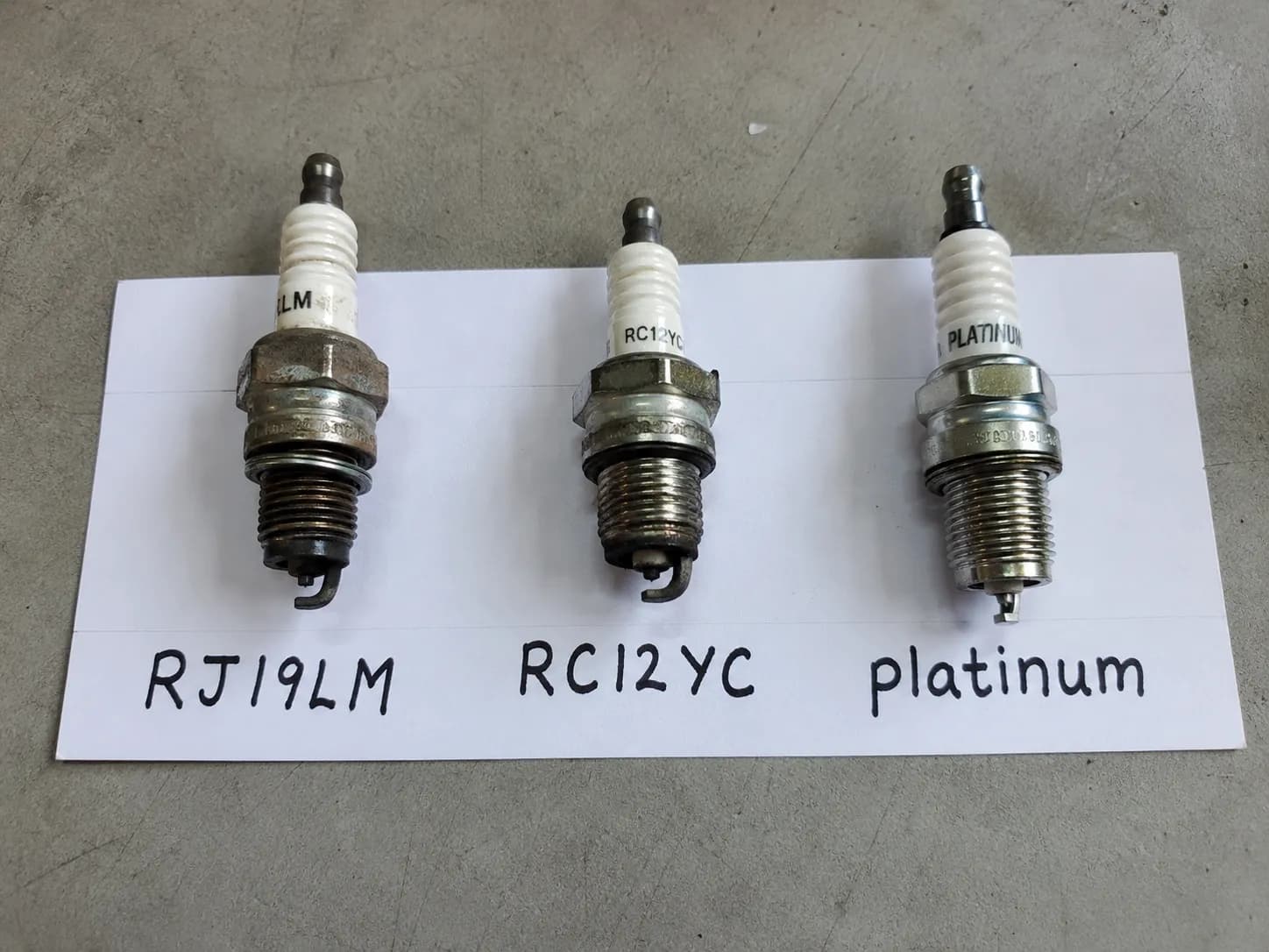 Three used Briggs & Stratton spark plugs labeled RJ19LM, RC12YC, and platinum, arranged side by side to compare thread length and electrode wear.