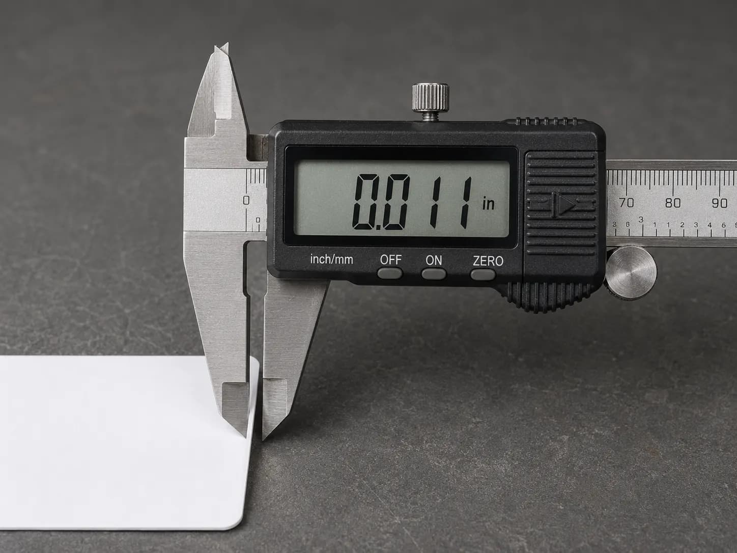 Digital calipers measuring a business card thickness at 0.011 inch on a workbench, demonstrating a simple thickness reference for lawn mower spark plug wire repair.