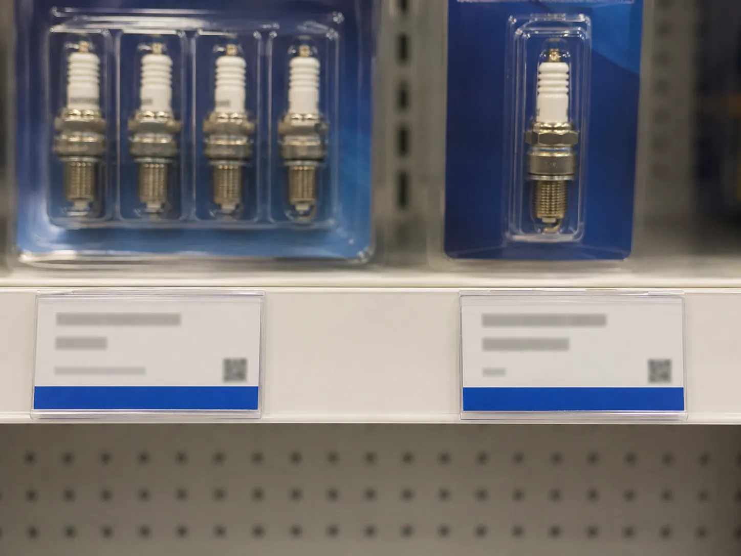 Close-up of adjacent retail shelf tags beneath a Champion four-pack spark plug and a Briggs Platinum single spark plug, showing their placement on the same store shelf rail.