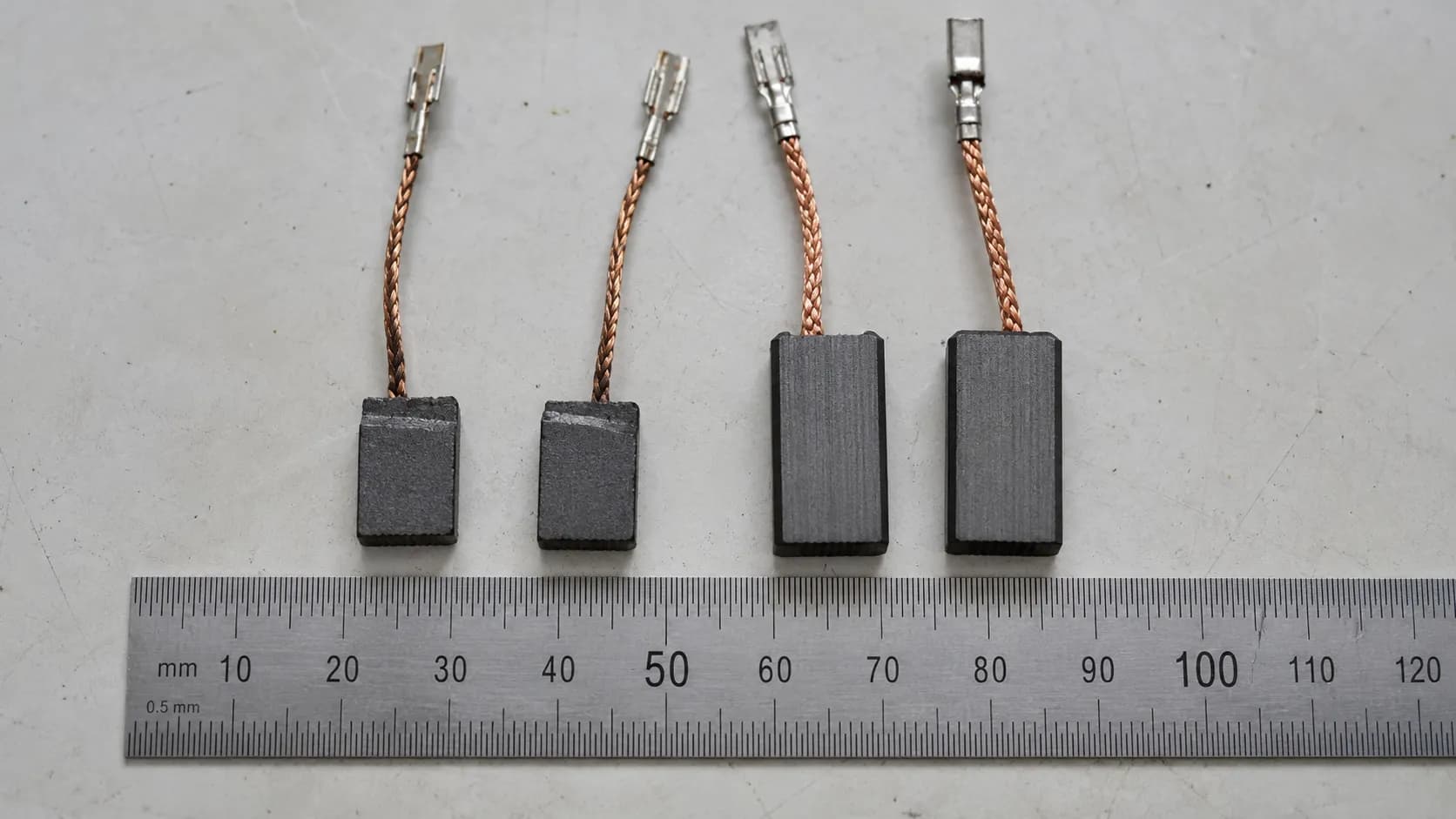 Worn and new carbon motor brushes from a corded lawn mower aligned beside a steel ruler, showing brush length wear after eight months compared with a new replacement pair.