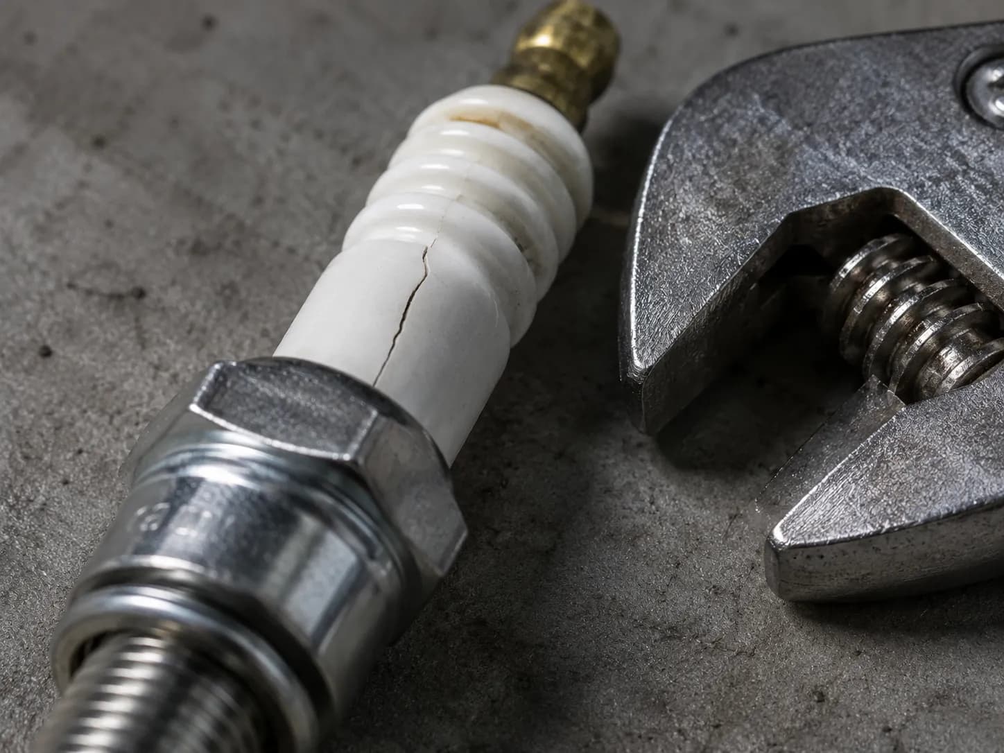 Close-up of a spark plug with a visible crack in the white ceramic insulator beside an adjustable wrench, illustrating how using the wrong tool can damage a spark plug during removal or installation.