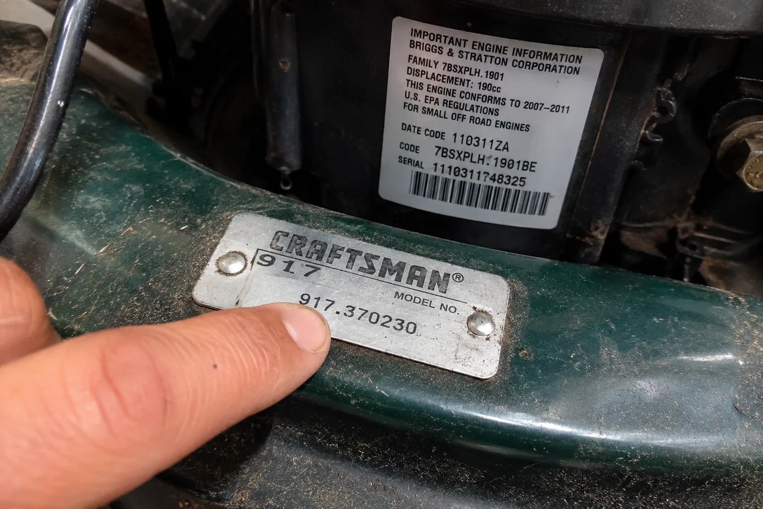 Close-up of a Craftsman mower model number tag on the deck with a finger pointing to the code, while the engine ID sticker is visible on the engine shroud in the background.