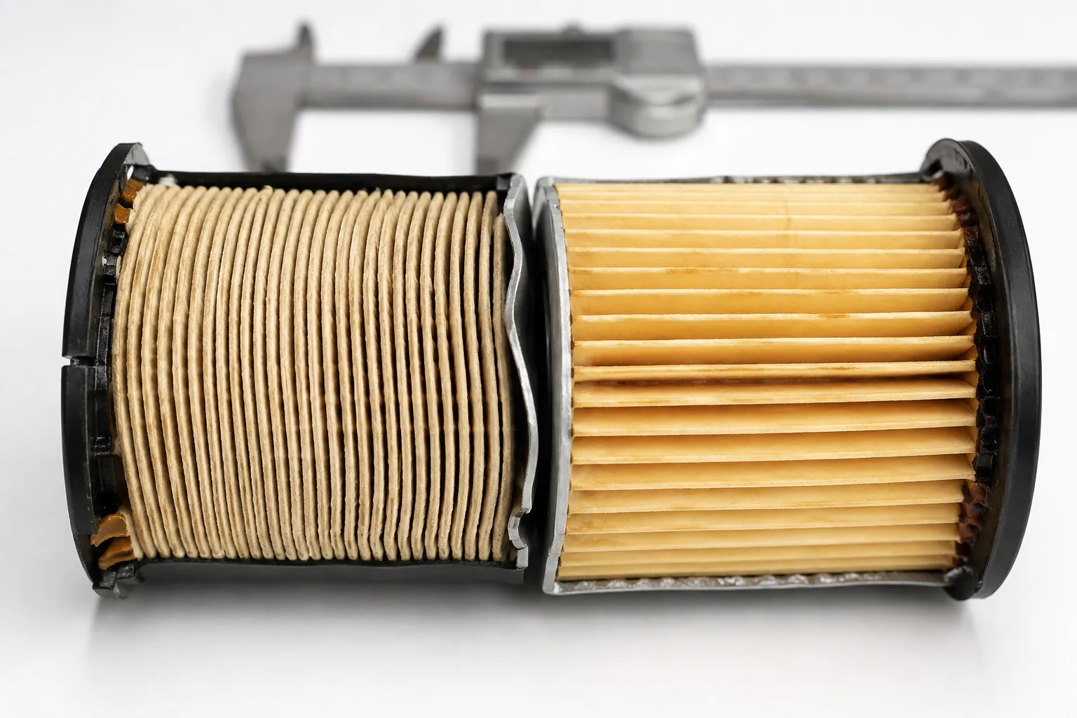 Two cut-open cylindrical oil filter elements placed side by side on a white surface, showing different pleat densities with digital calipers blurred in the background for comparison.