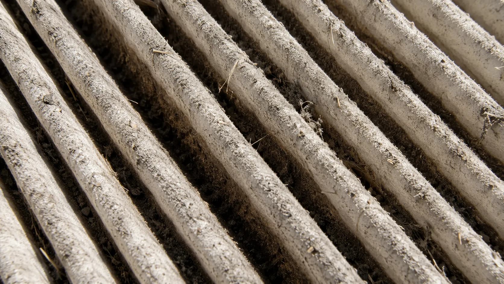 Macro close-up of a dirty paper air filter showing loose dust and grass chaff on pleat ridges with dark embedded staining deep between the pleats under raking side light.