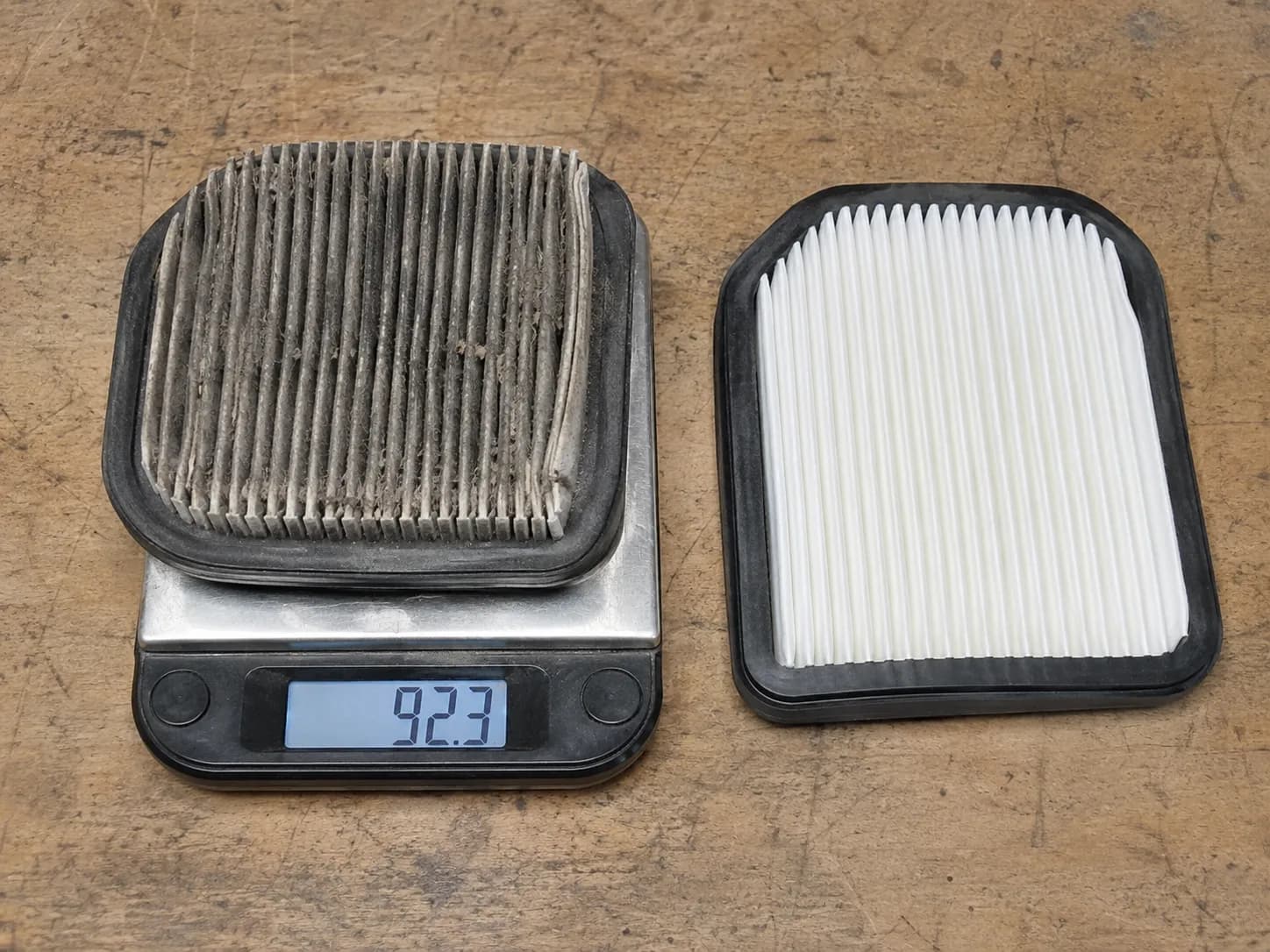 Dirty paper air filter on a digital scale beside a new replacement filter, showing the weight comparison between a dust-loaded used element and a clean paper air filter on a workshop bench.