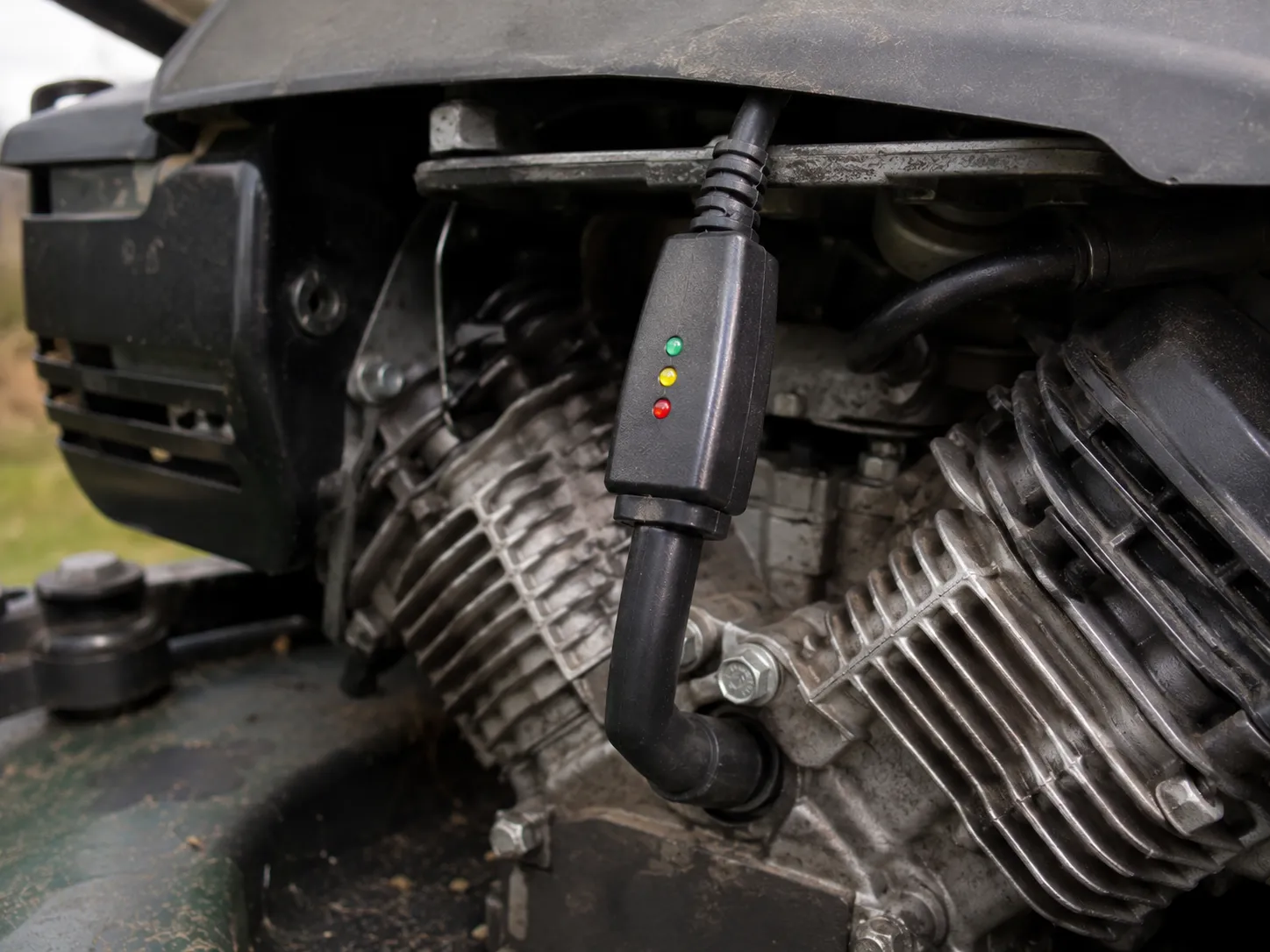 Electronic spark checker clipped to the spark plug lead on a Kawasaki V-twin mower engine with the spark plug left installed, showing a non-invasive ignition spark testing setup.