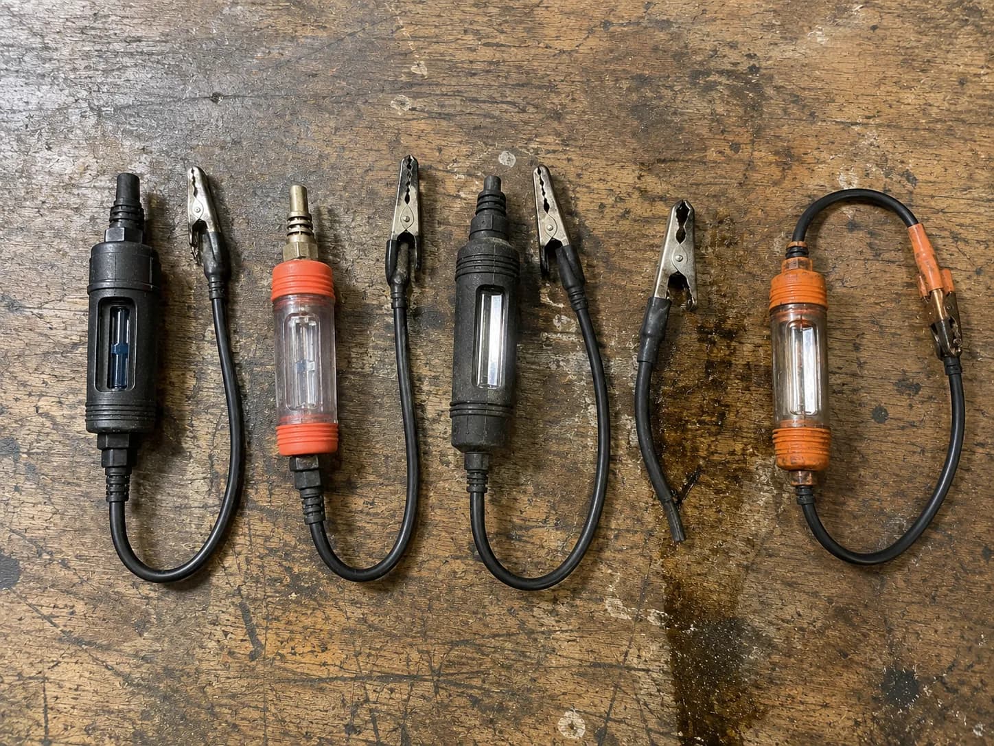 Five inline spark testers after durability testing, showing worn rubber boots, scuffed housings, a sprung ground clip and oil marks on a wooden workbench.