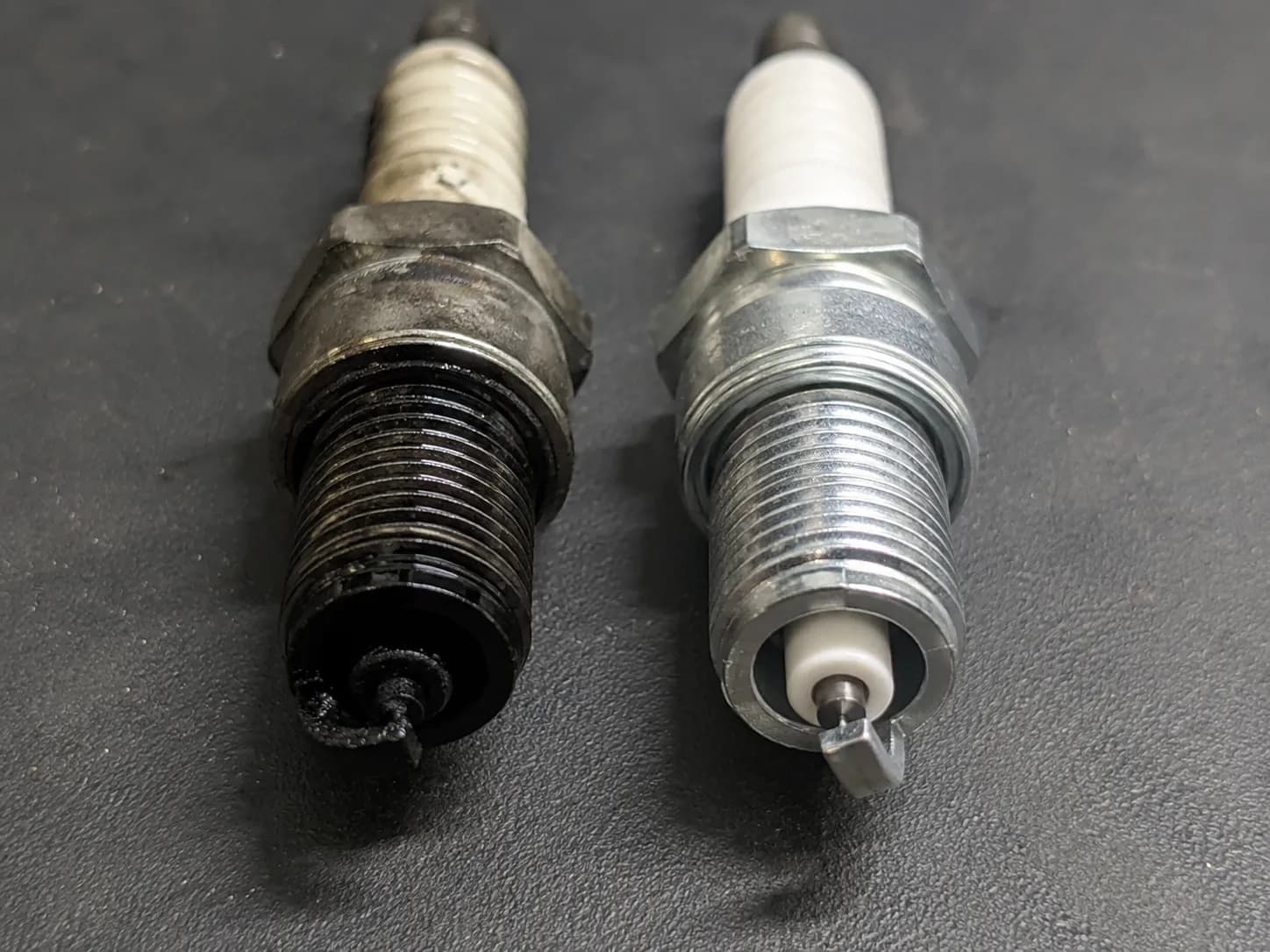 Wet, black fuel-fouled spark plug beside a clean replacement after a flooded small engine cylinder, showing the difference in plug condition.